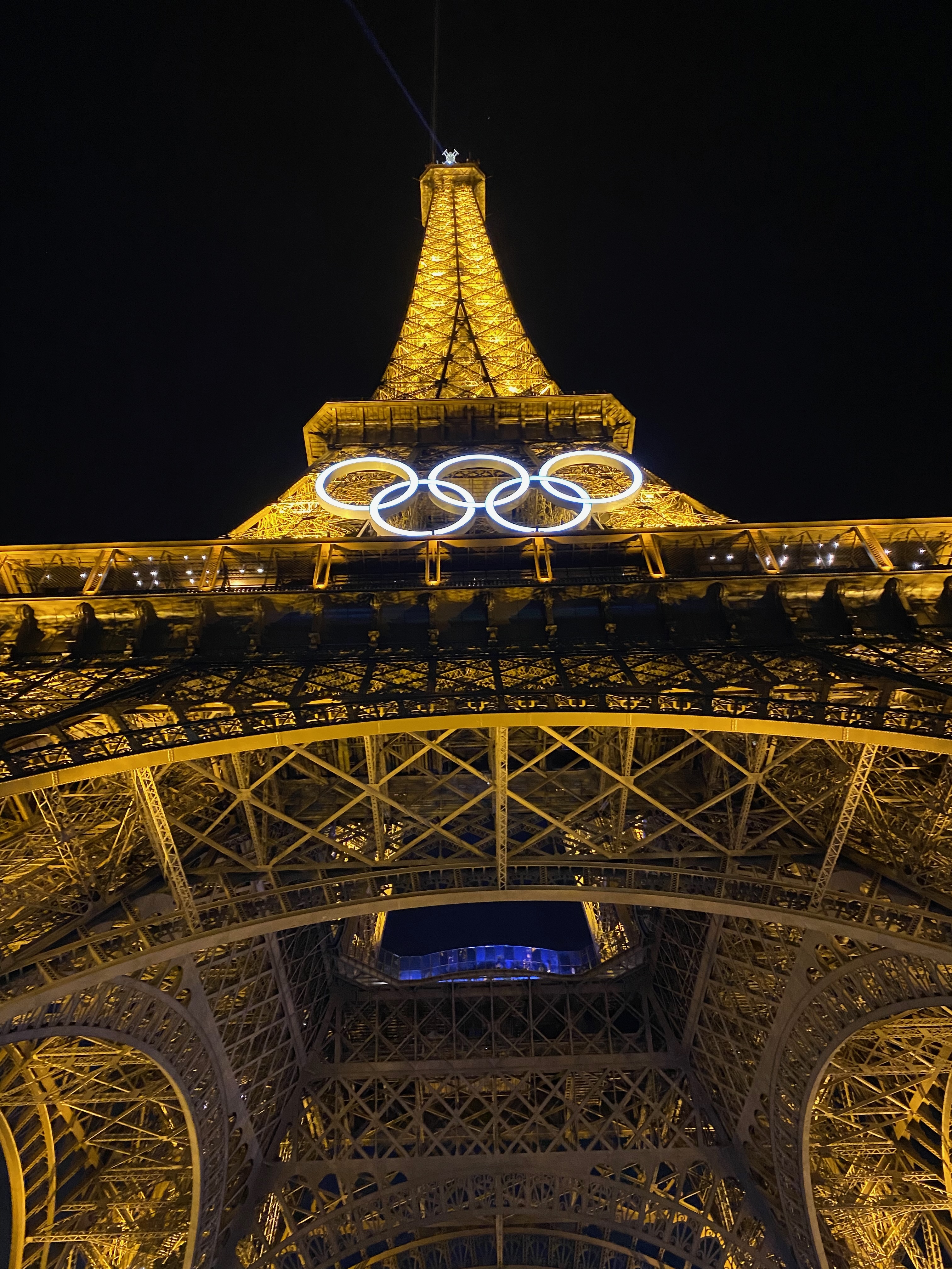 View from the bottom of the Eiffel Tower of the structure lit up at night with the Olympic rings hanging in front