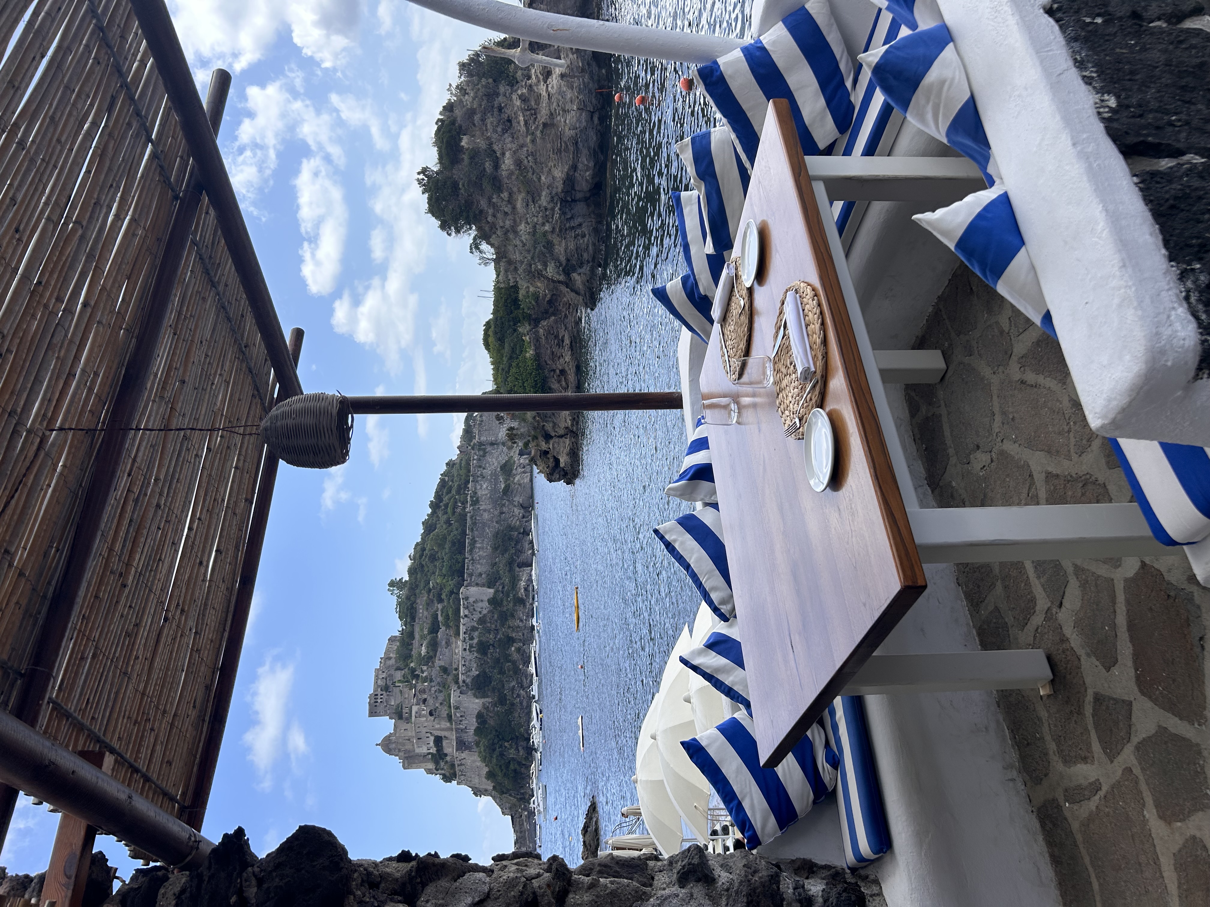 View of a wooden table set with plates and white and blue striped pillows by the sea