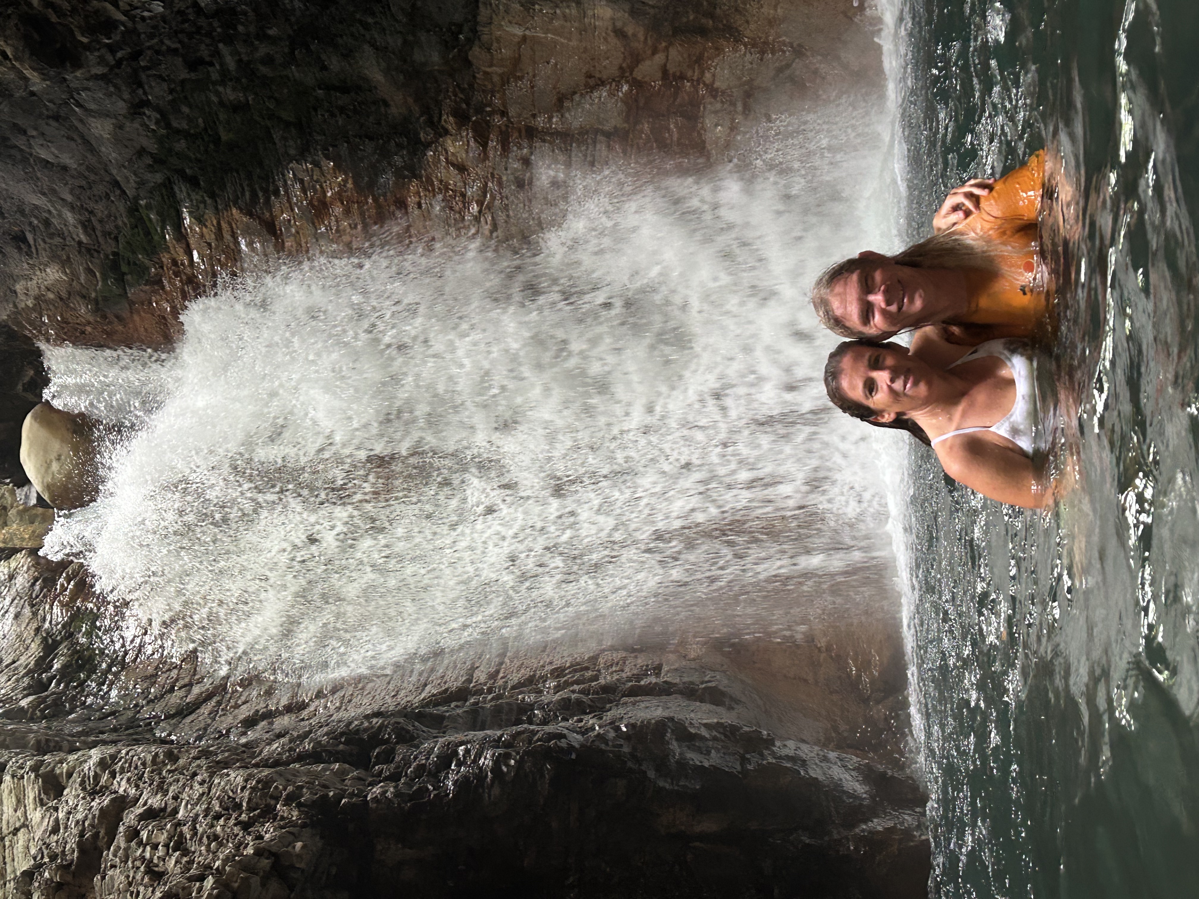 A view of two people posing in a larger pool of fresh water with a waterfall in the distance on a sunny day.