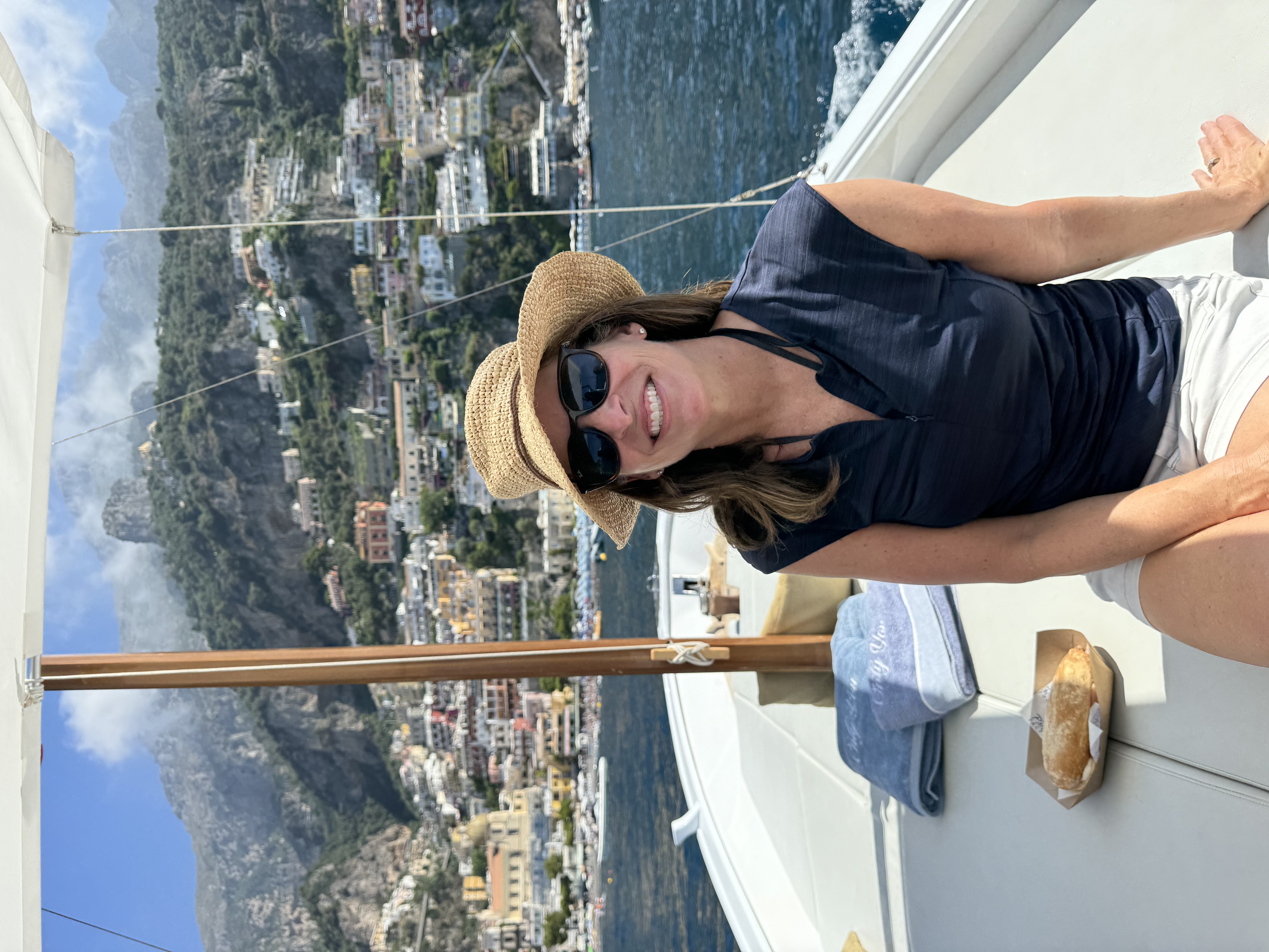 Advisor in a sun hat and sunglasses sitting on the deck of a boat with a coastal town on a hill in the background