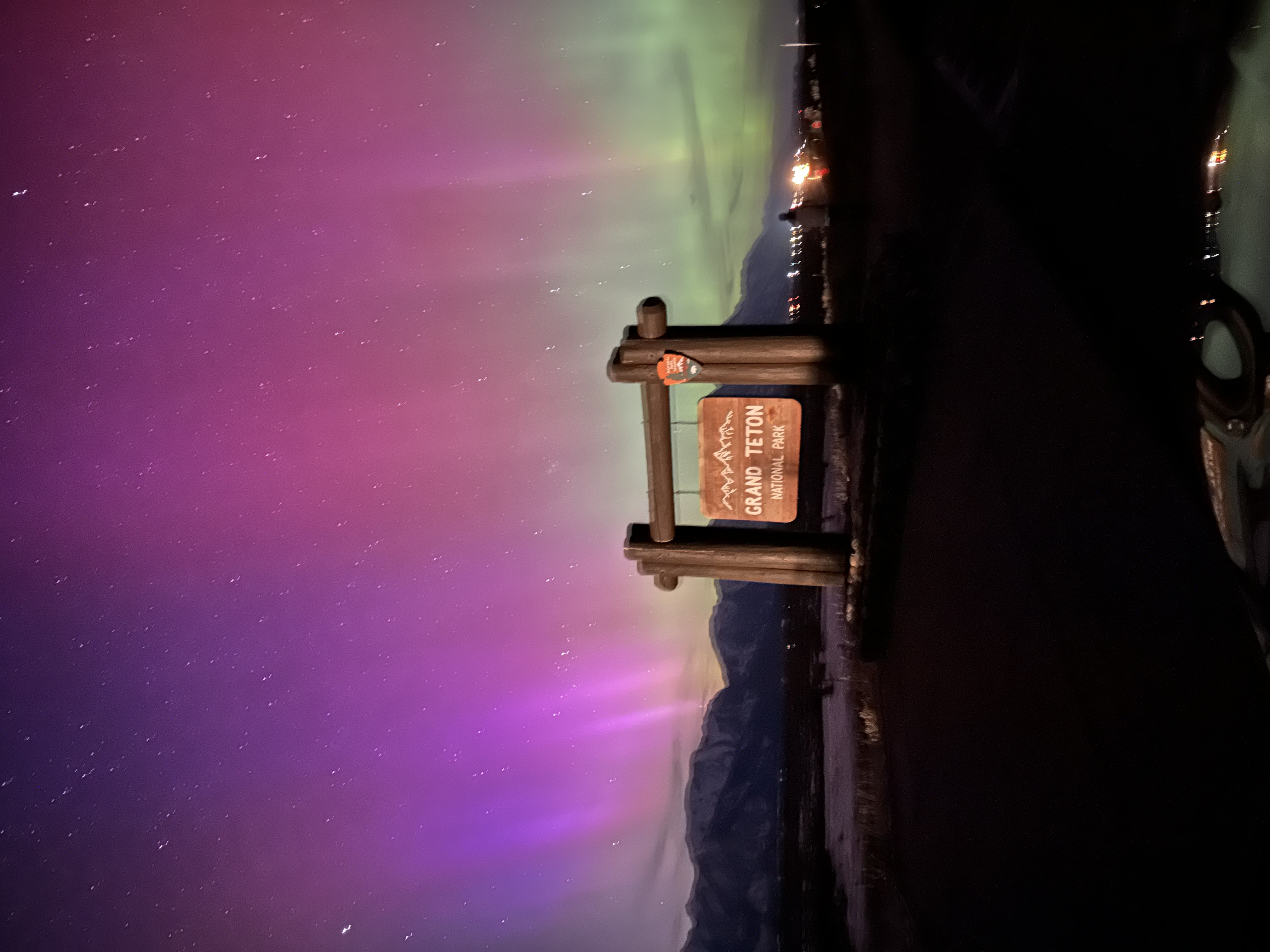 View of bright pink and green northern lights over a hanging wooden sign in the distance