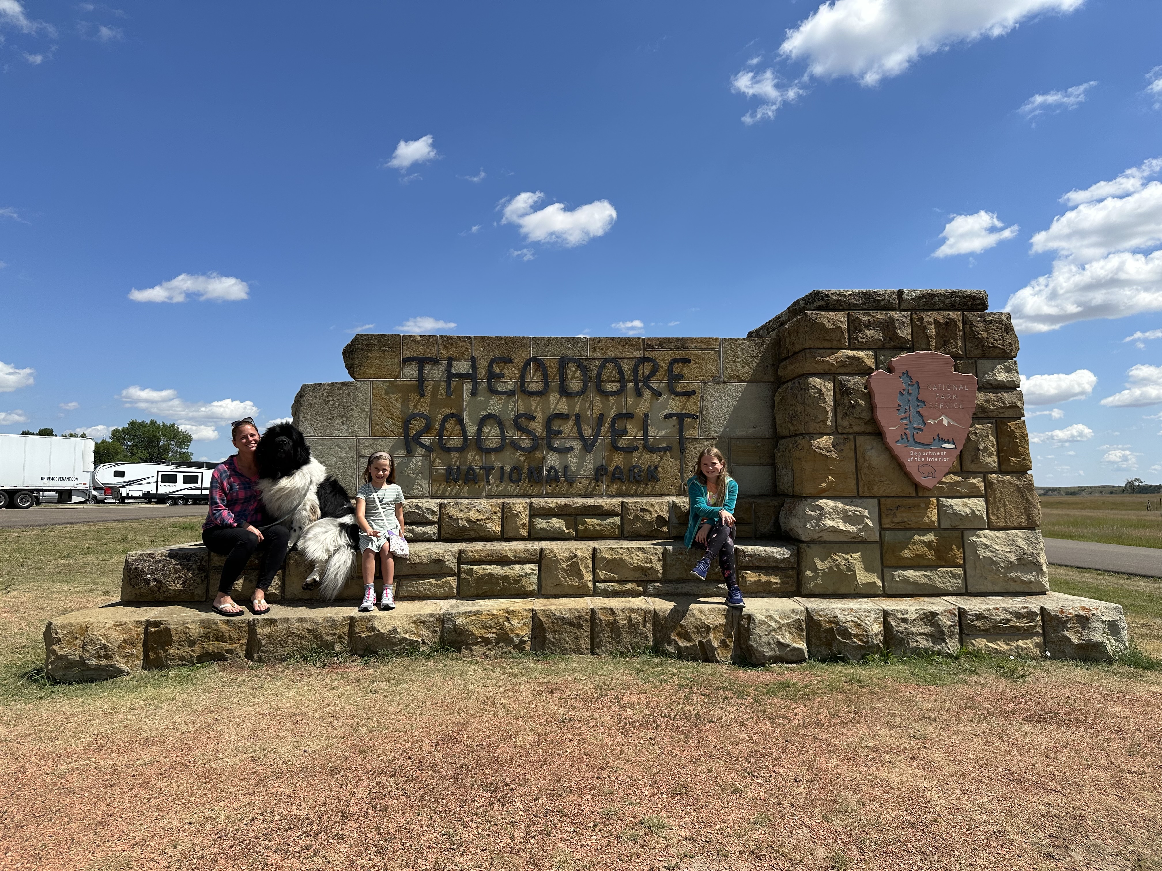 Advisor and family sitting in front of the sign for Theodore Roosevelt National Park on a sunny day