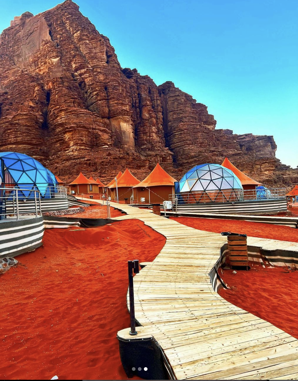 View of a wooden path over red sand with dome-shaped accommodations on either side