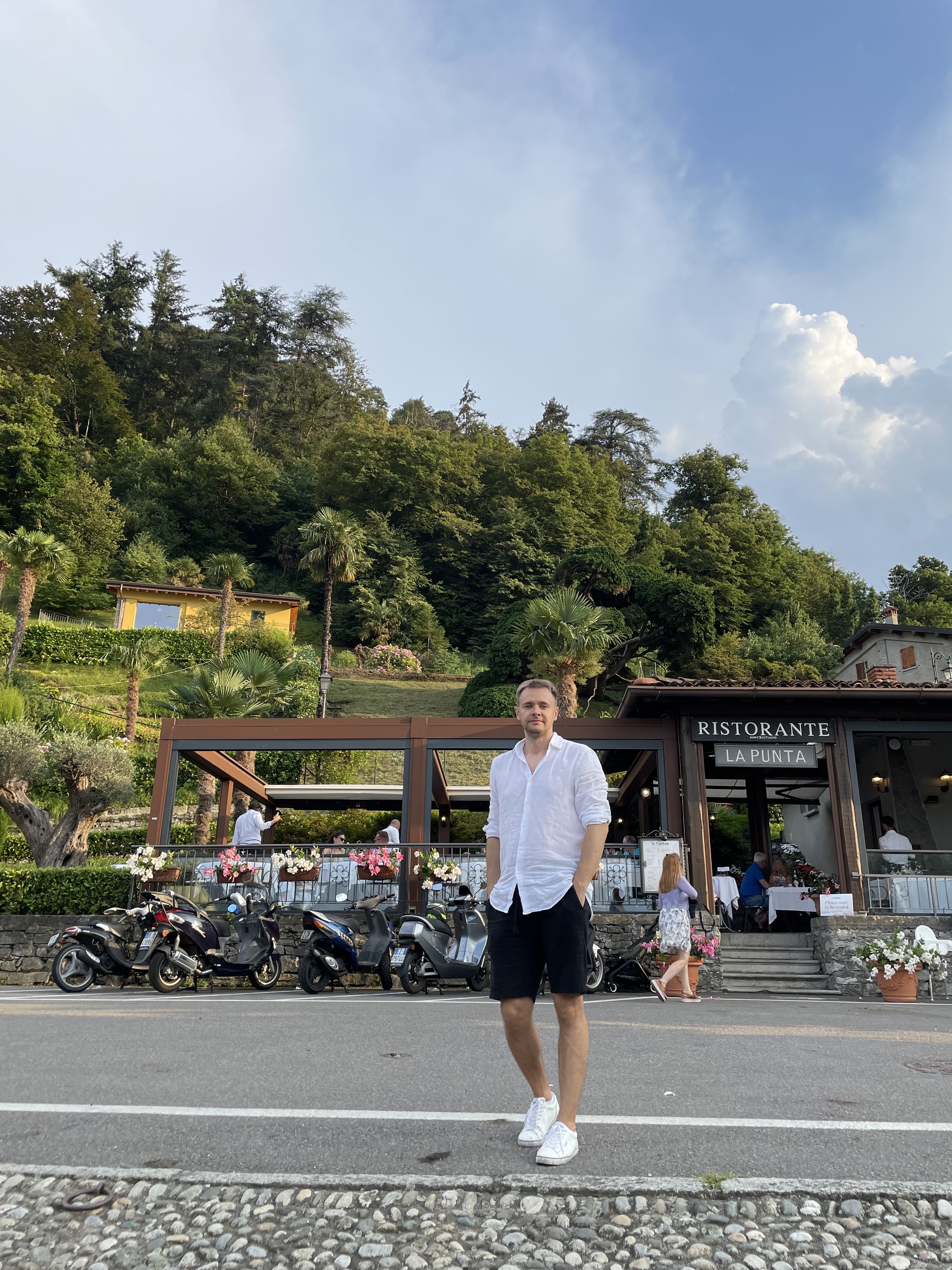 A man posing in front of a restaurant outside surrounded by lush trees and parked motorbikes.