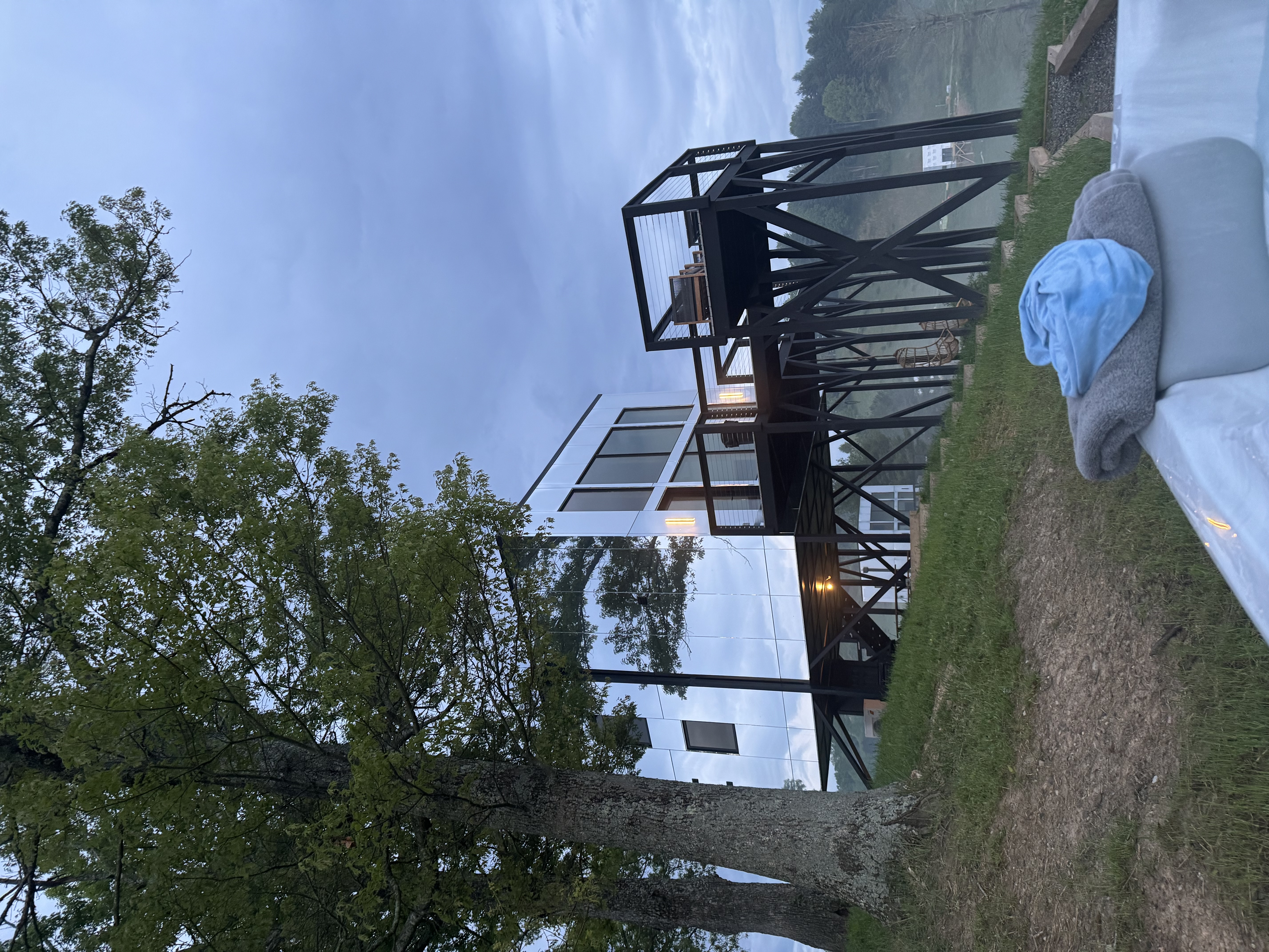 A view looking at a home with mirrored siding on a hill with a deck on stilts on an overcast day.
