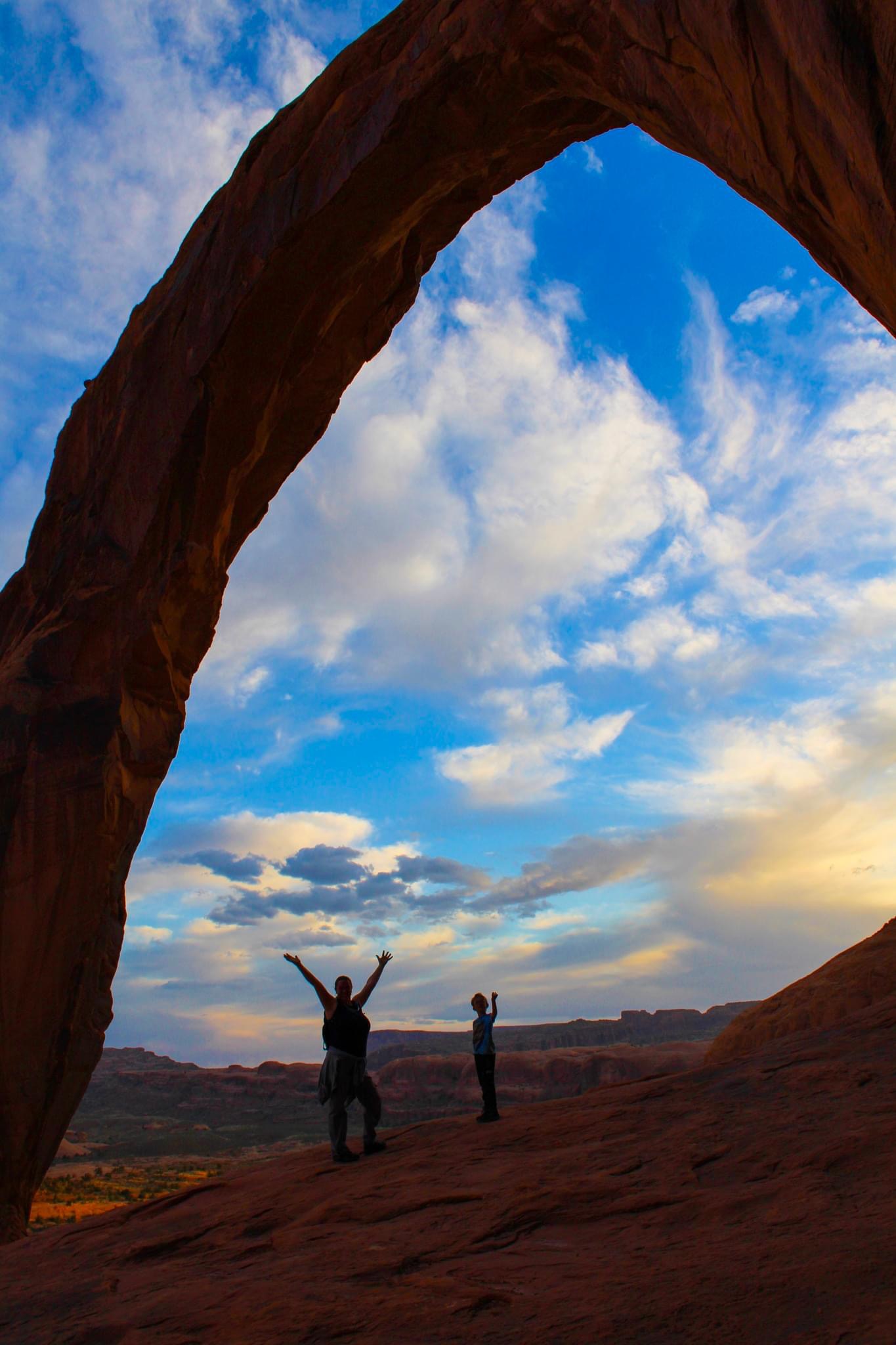 A view through a rock formation arch with their hands in the air in a desert landscape and a partly cloudy sky above.