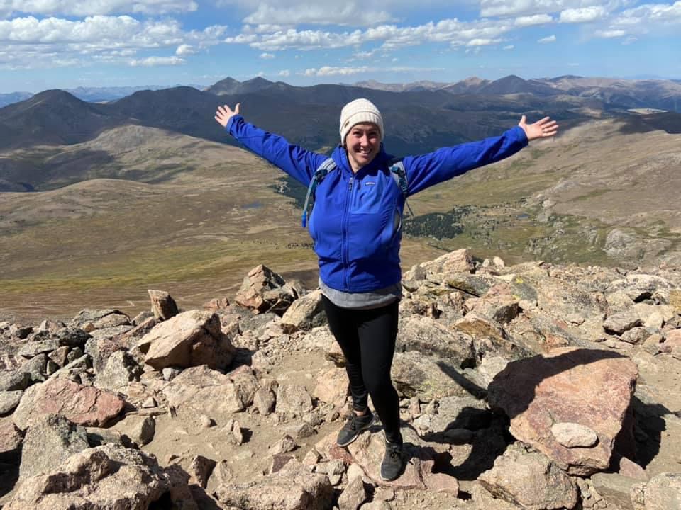 Advisor standing with open arms on a rocky mountainside with a mountain range behind her during the day.