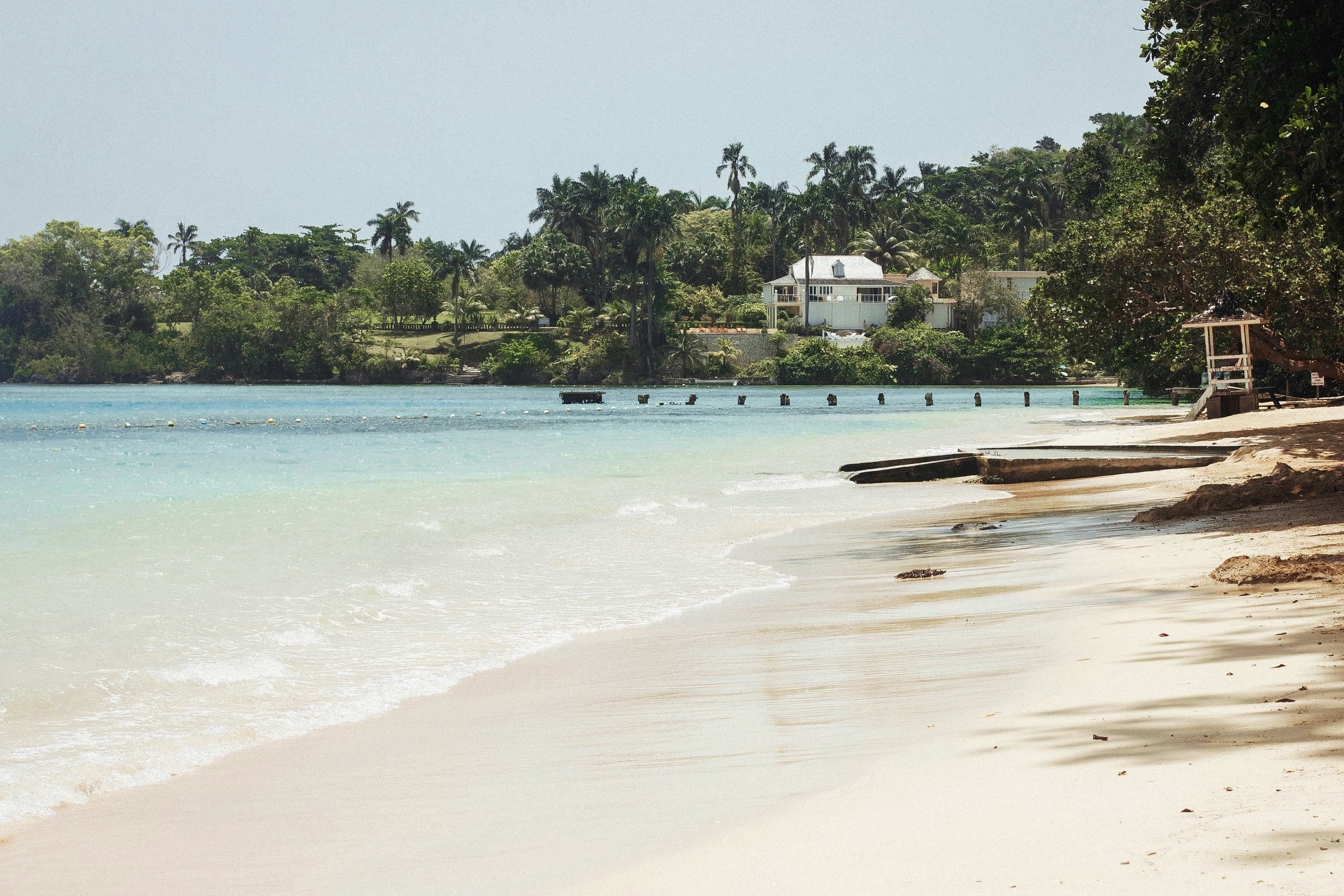A view of white sand beach and a doc leading out to the water with palm trees and a white house on the beach.