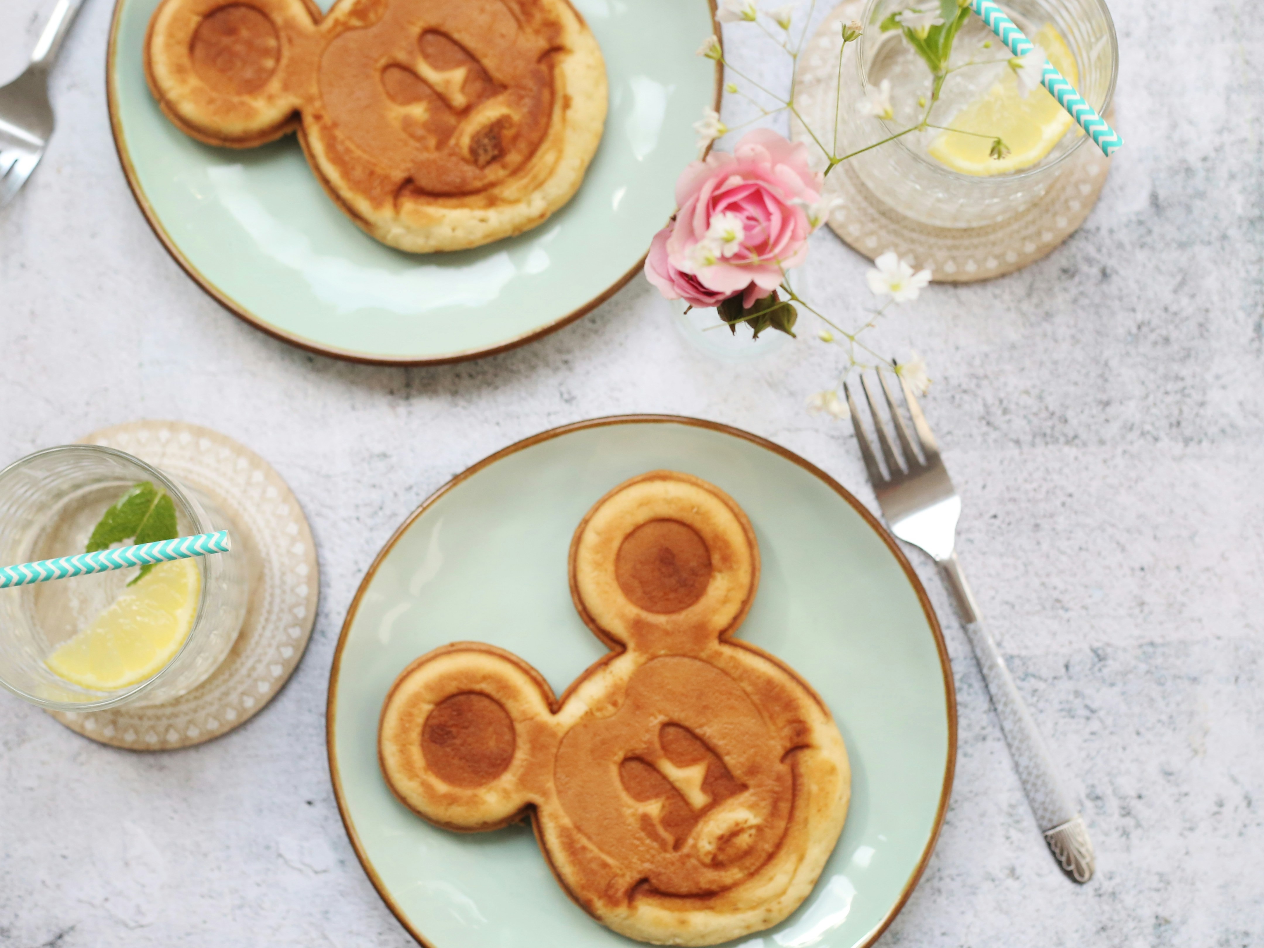 Two plates with pancakes shaped like a cartoon character’s face, accompanied by drinks with lemon slices and a pink flower.