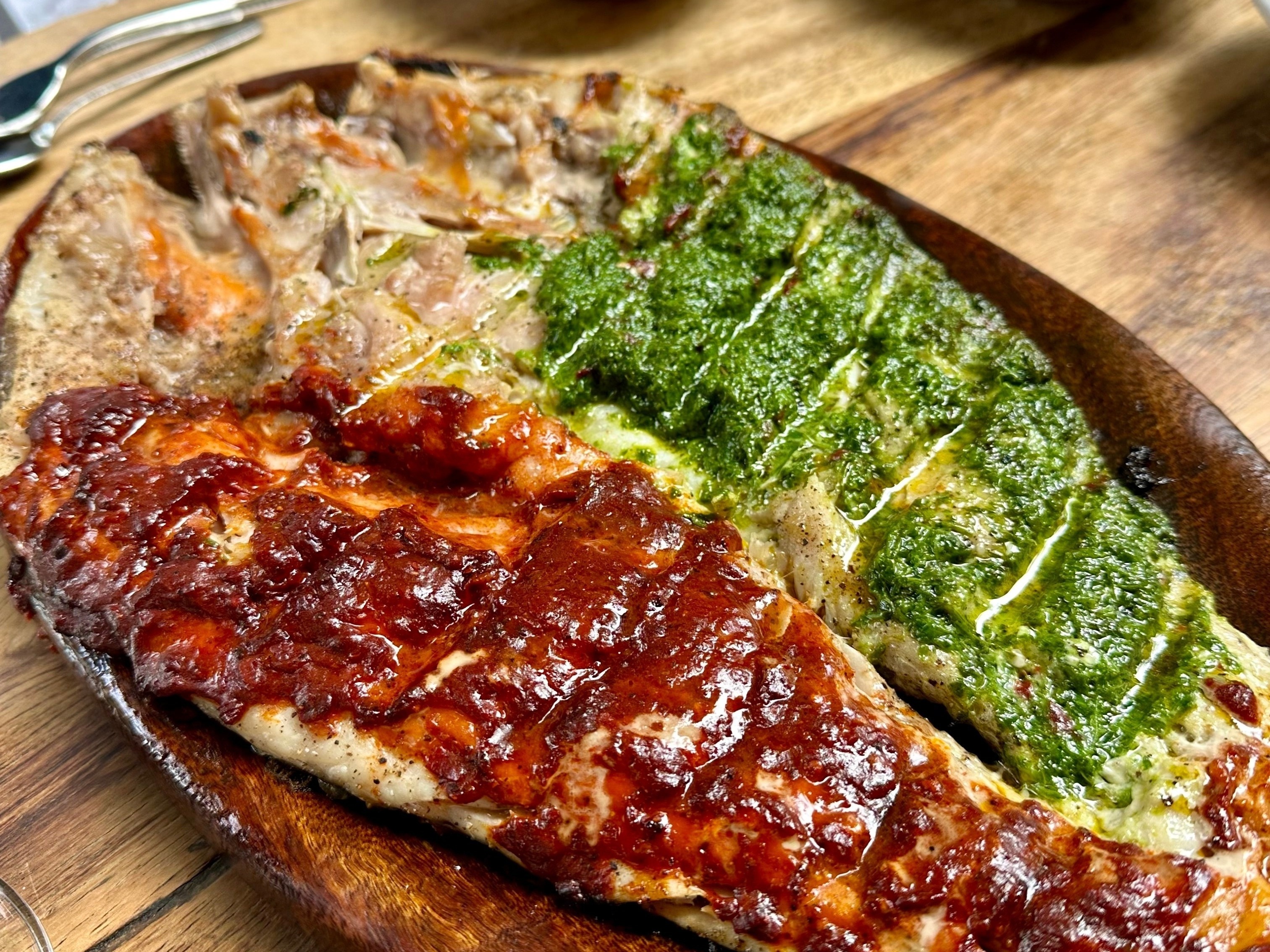 Meat covered in red and green sauces, sitting on a plate on a wooden table