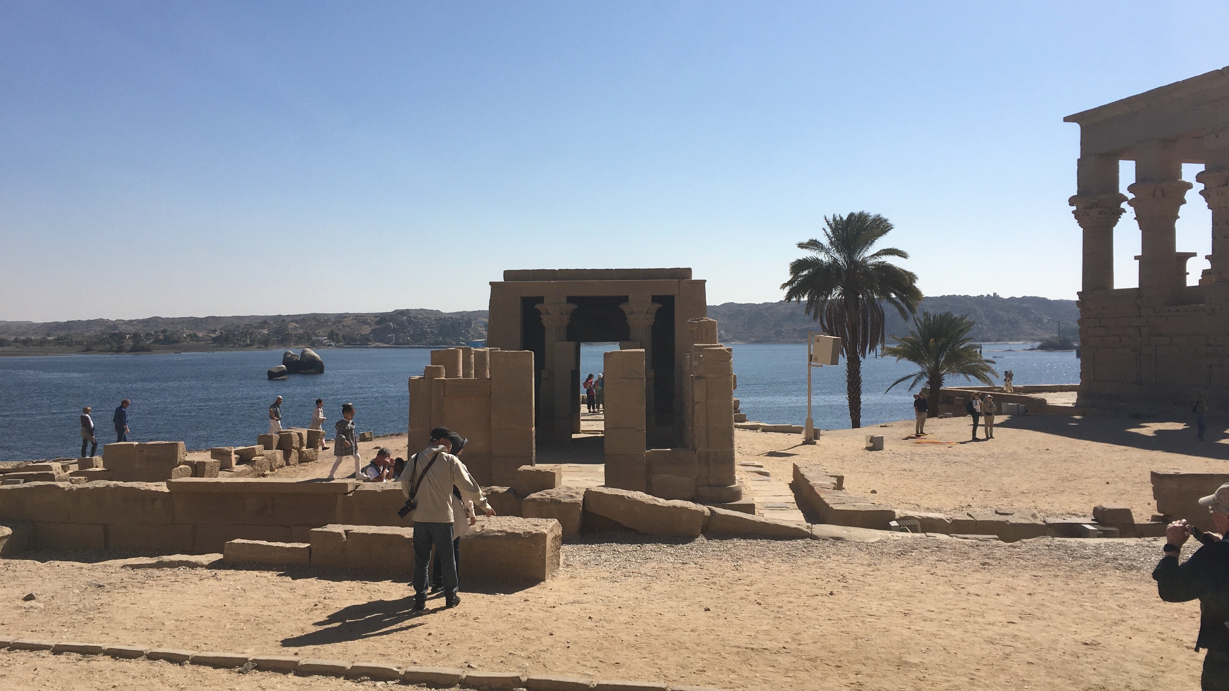 An ancient Egyptian temple complex with tourists, set against the backdrop of the Nile River and a clear blue sky.