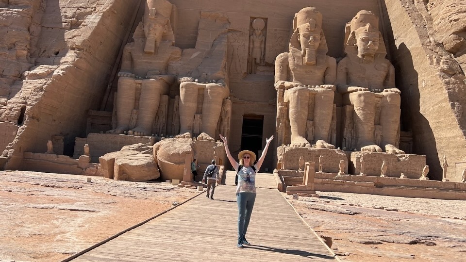A person with raised arms stands in front of the Great Temple of Abu Simbel, with its four colossal statues and entrance.