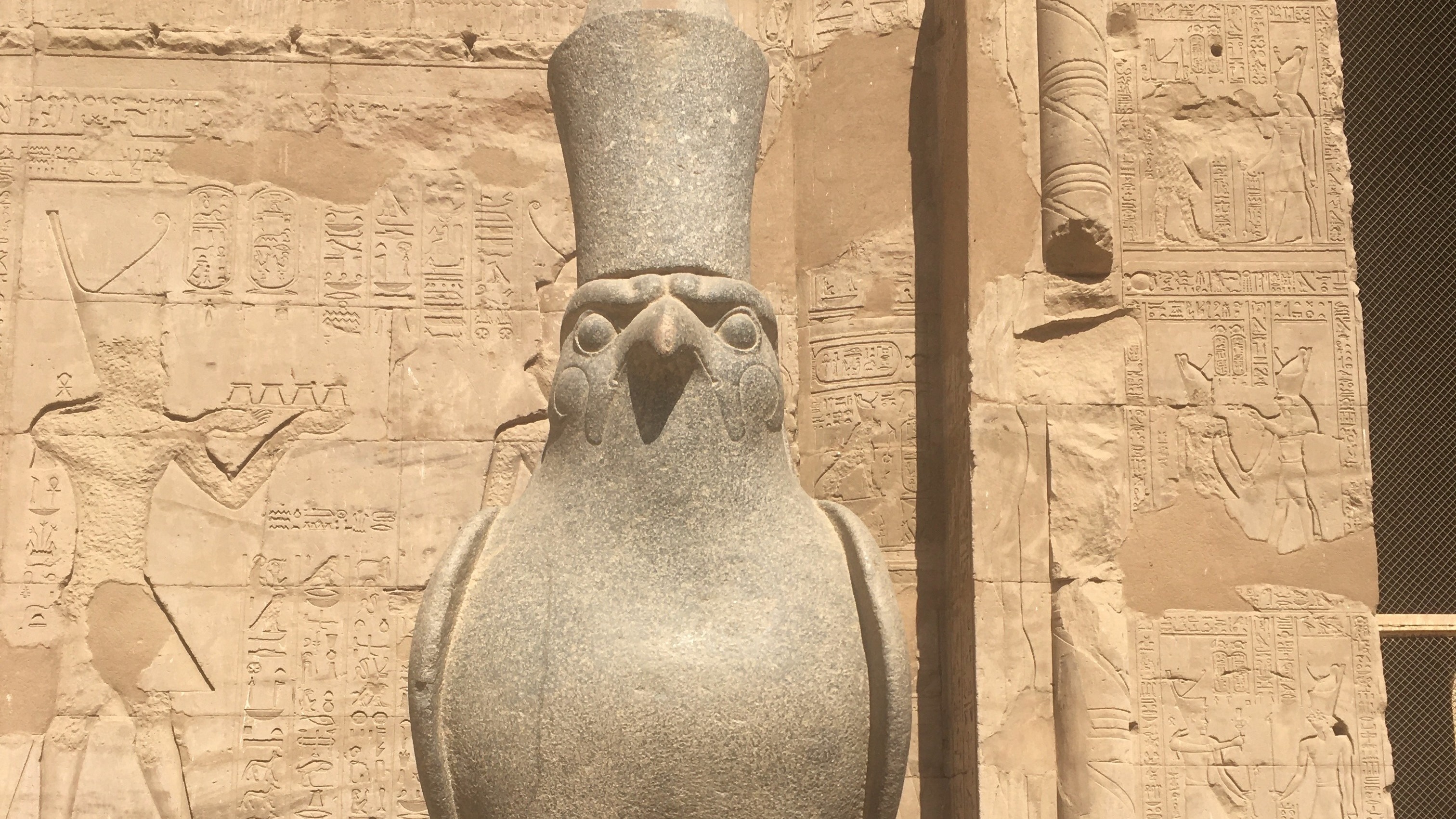 There is a large stone statue of a bird with a human figure carved on the wall behind it, possibly an ancient Egyptian artifact.