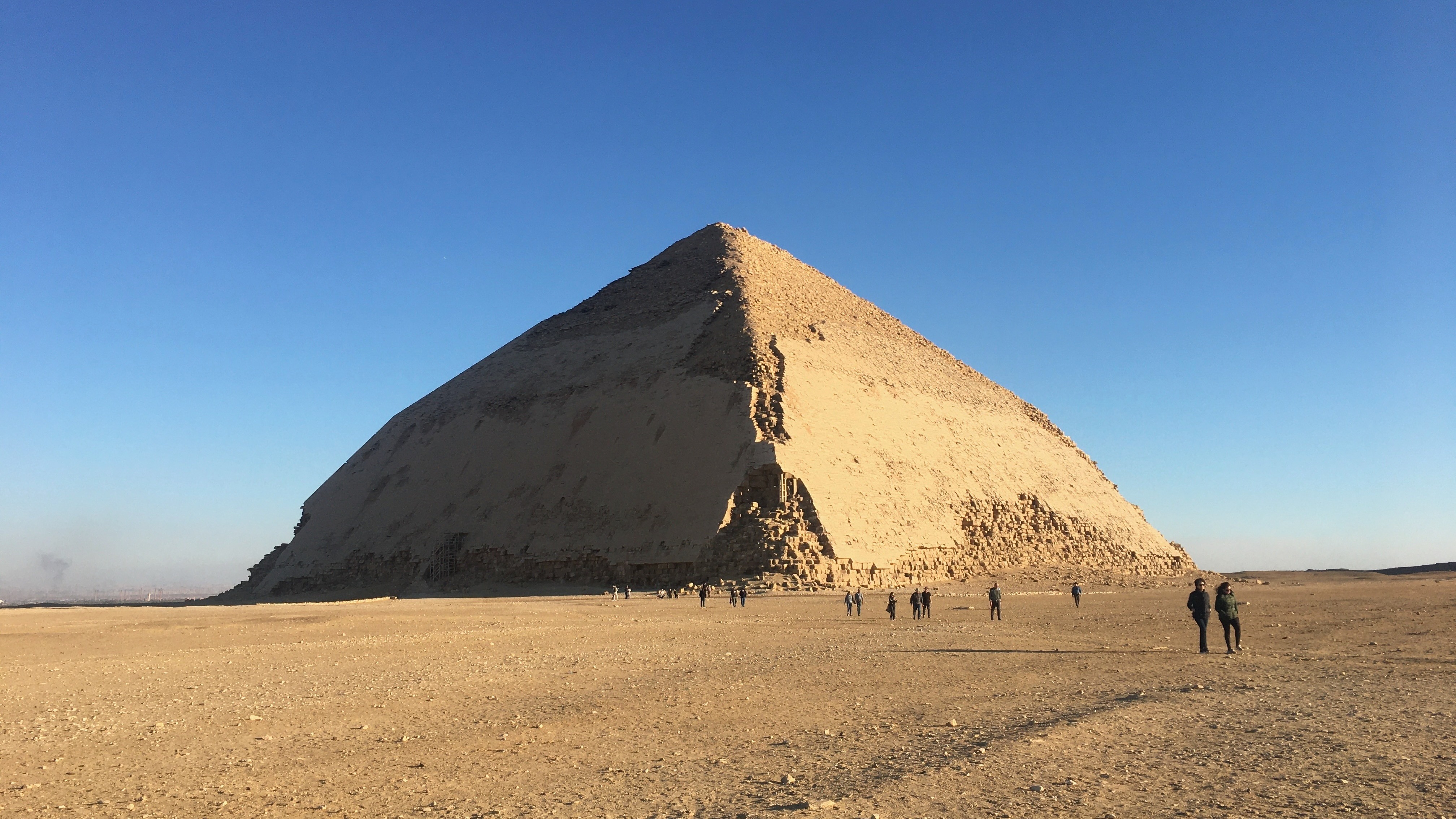 The Bent Pyramid in Dahshur, Egypt, under a clear blue sky with several people near its base, highlighting its unique architectural structure.