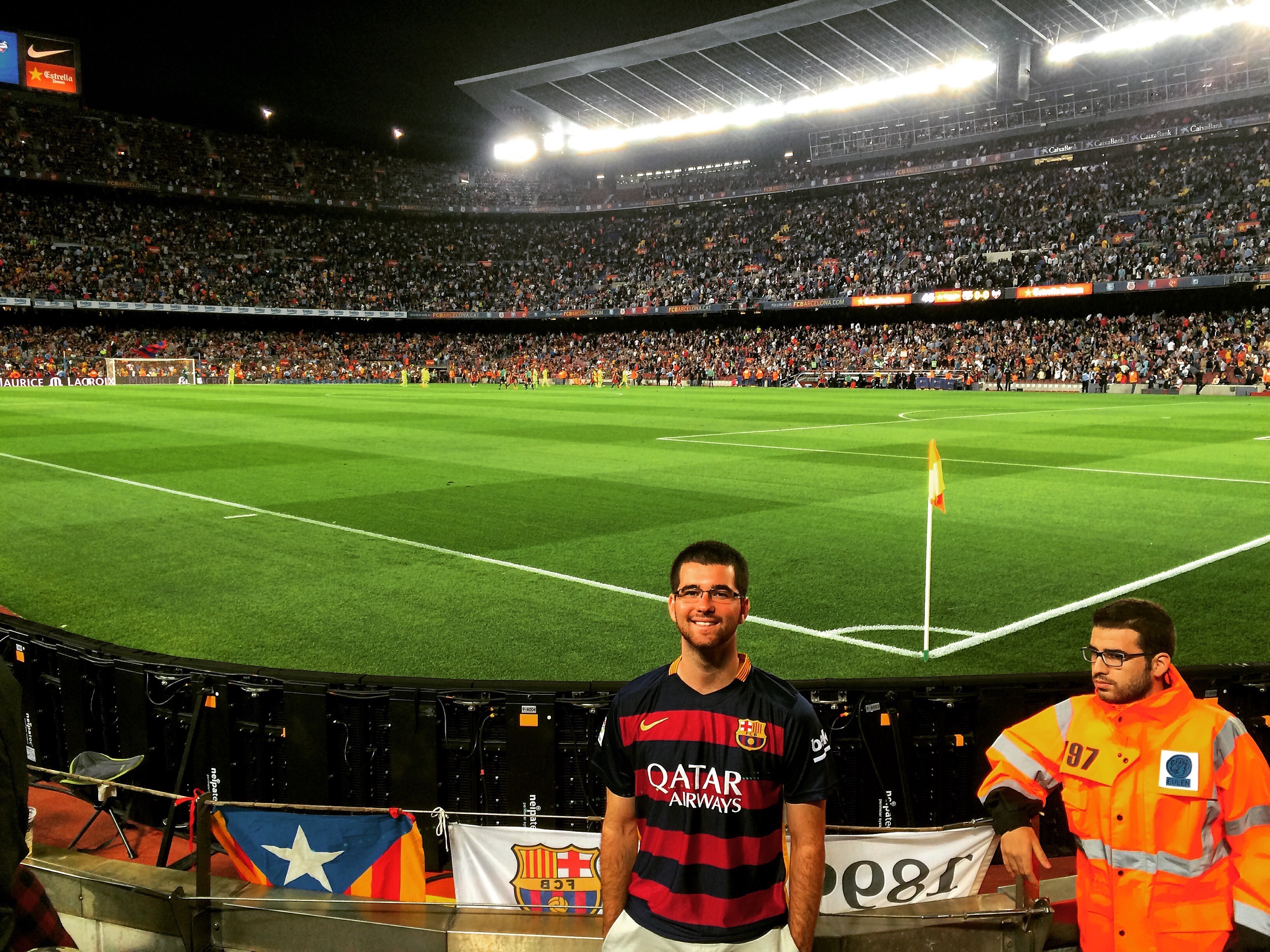 A person smiling for a photograph while standing in front of a soccer field in a stadium filled with people
