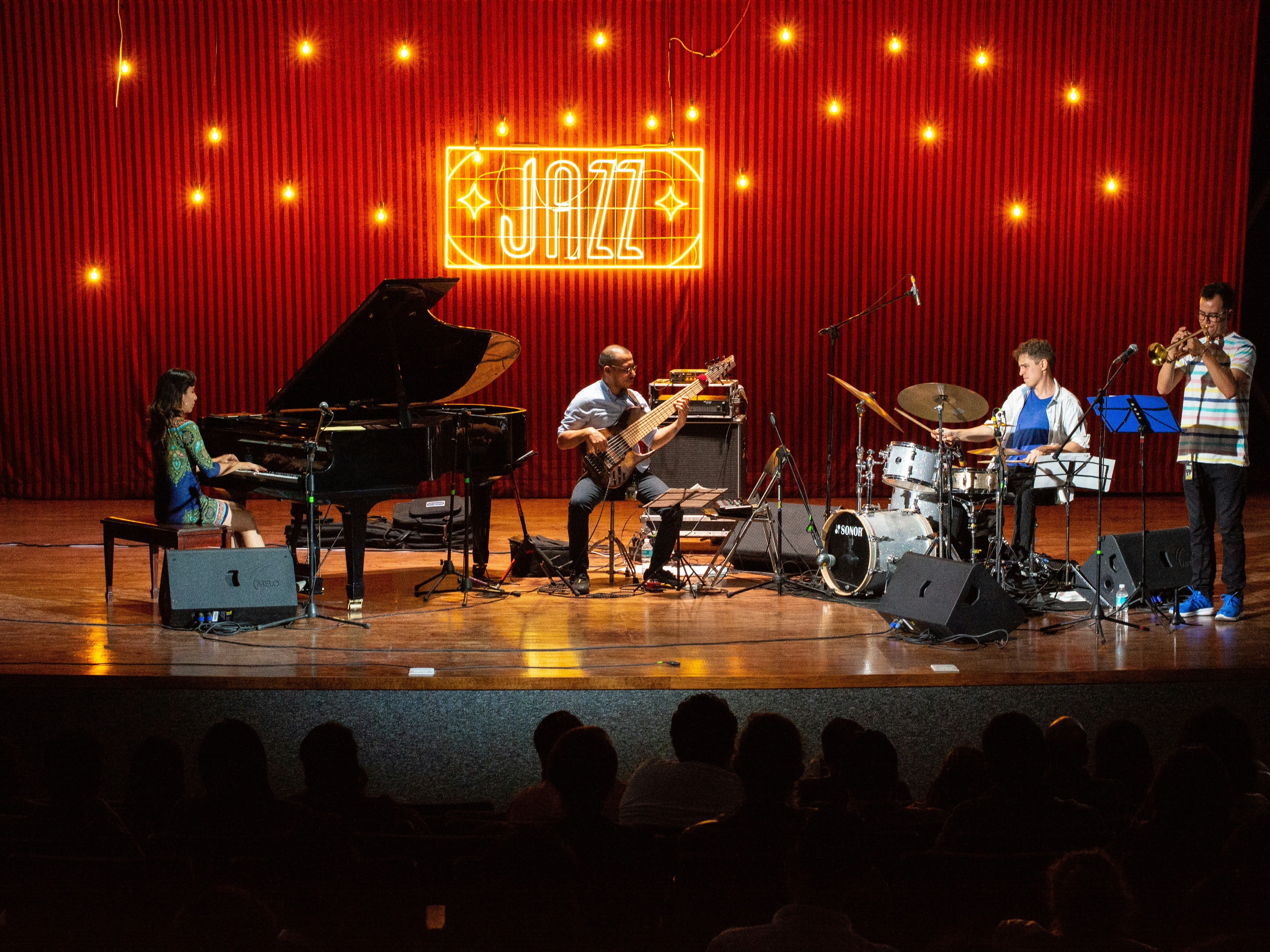 A band on stage in front of a red curtain and a light up sign reading "JAZZ"