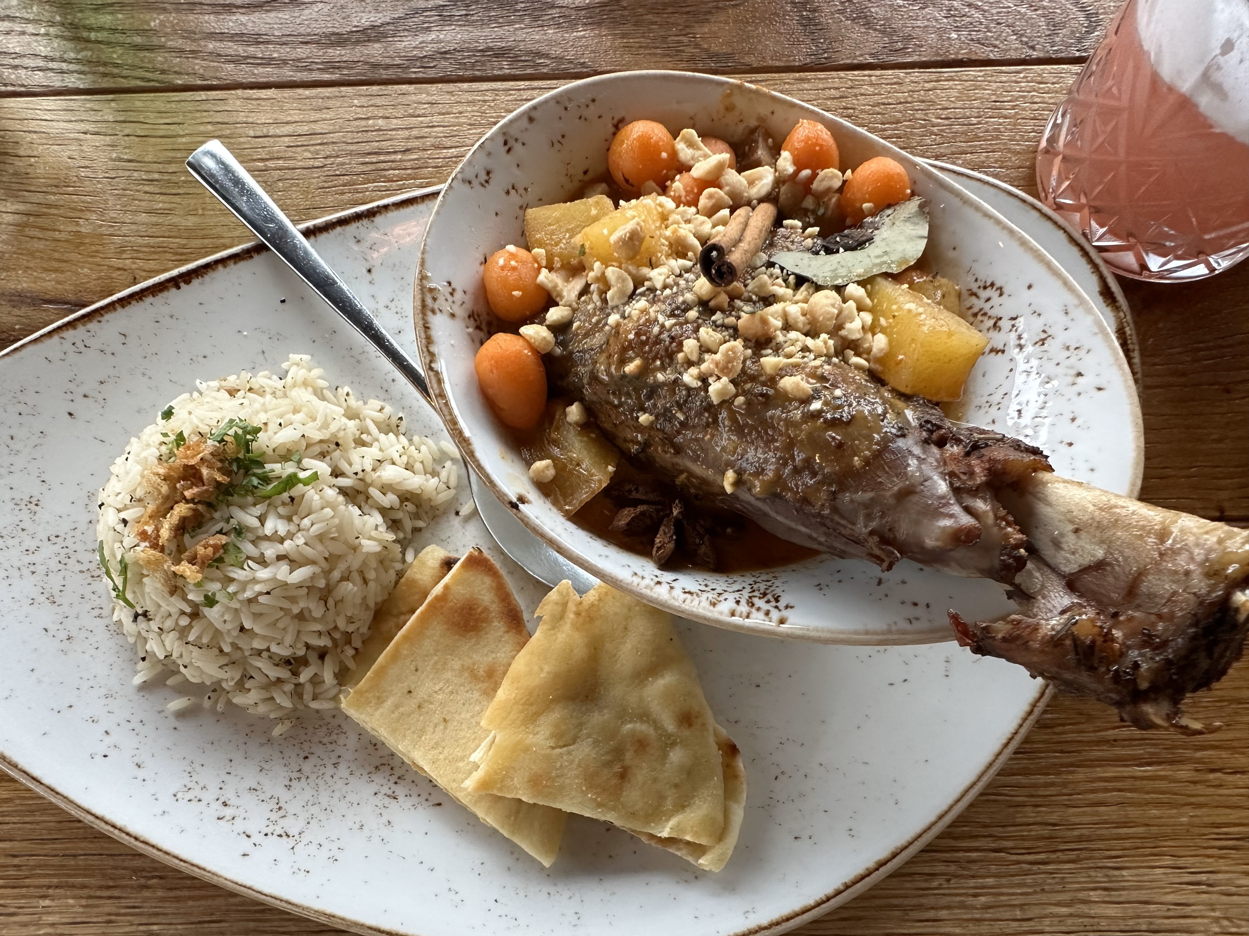 The image shows a plate with a large meat portion, rice, flatbread, and a mix of vegetables and nuts on a wooden table.