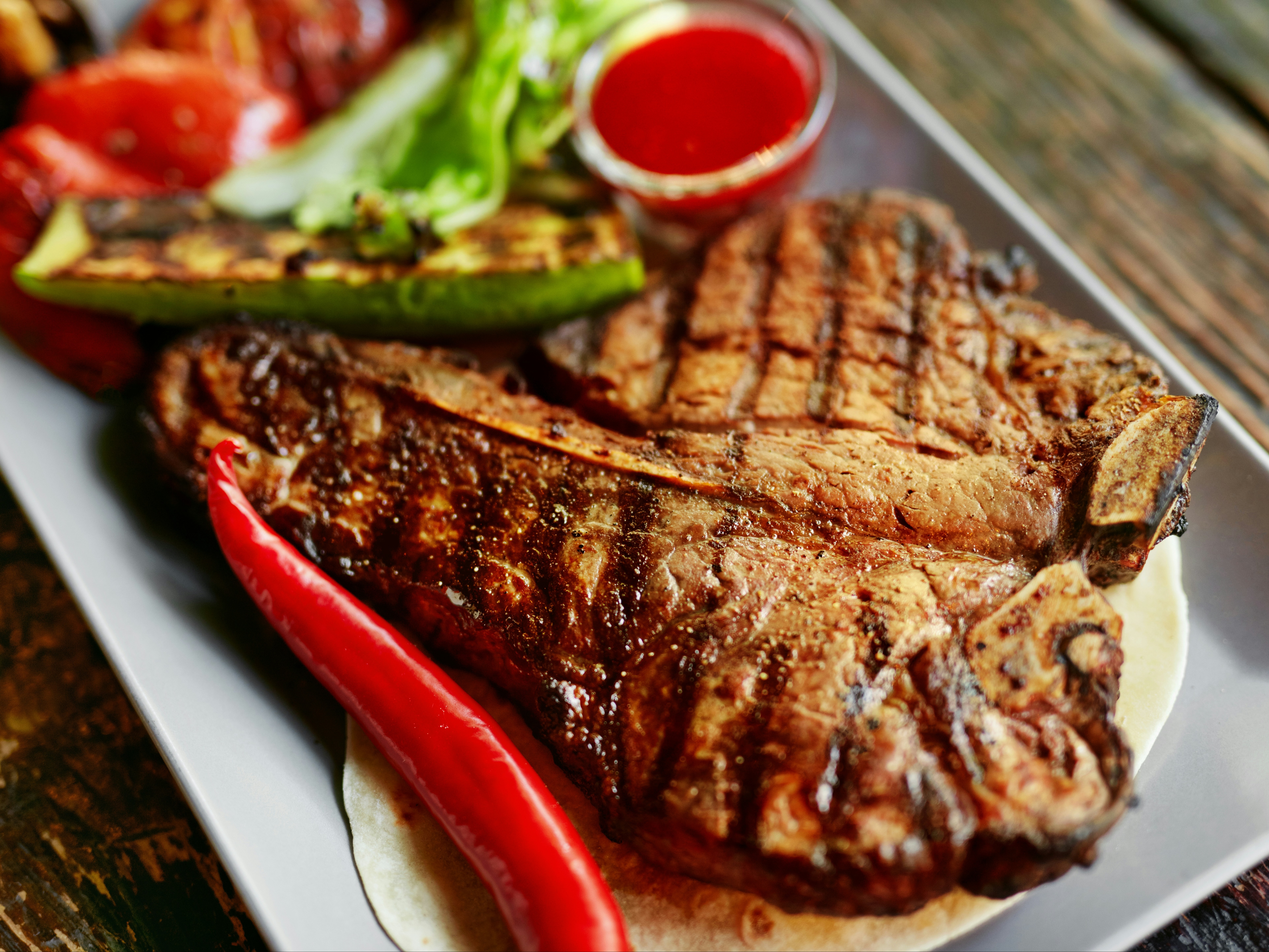 A grilled steak with char marks, accompanied by grilled vegetables and a sauce, on a wooden table.