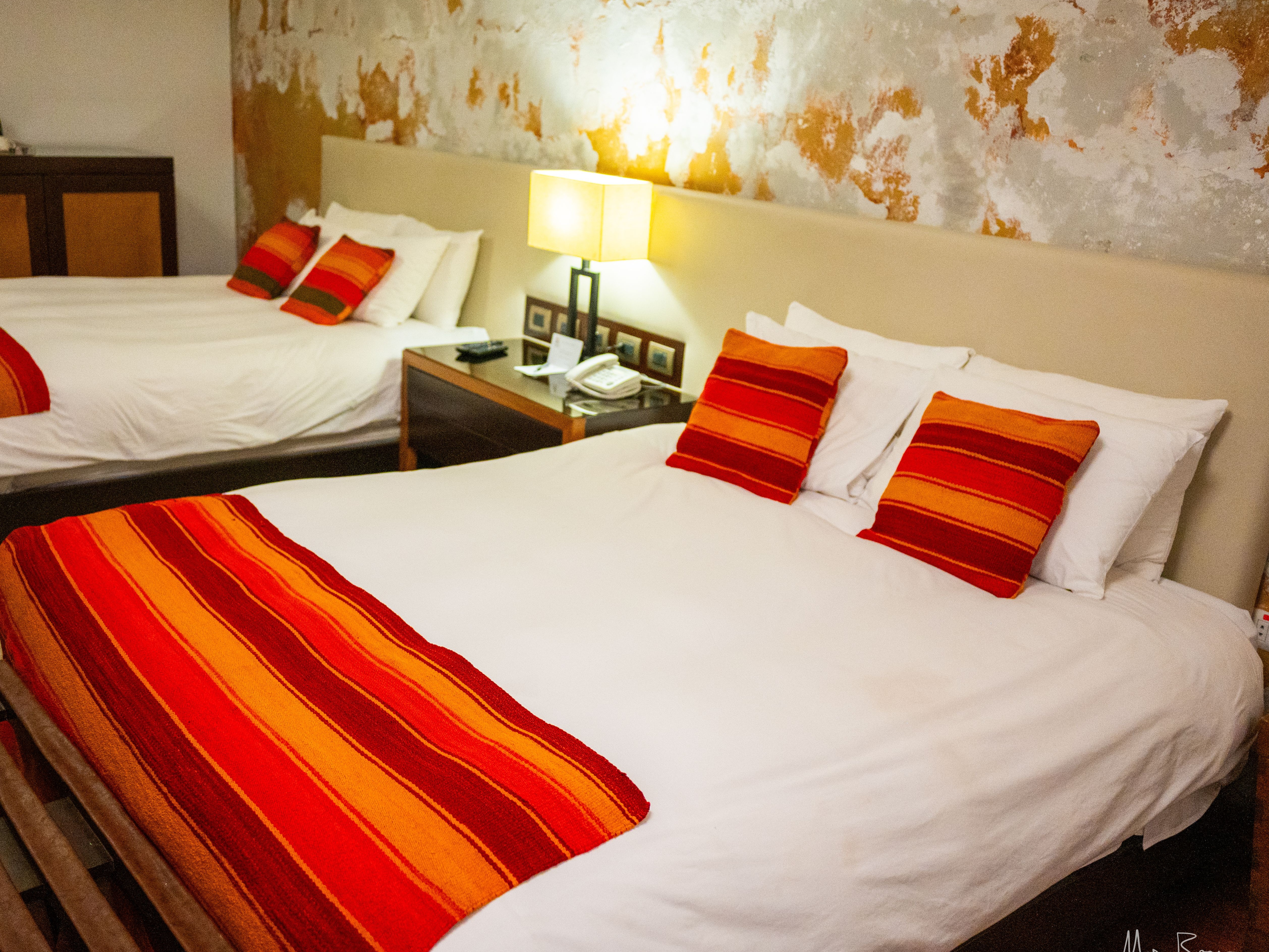 A well-lit hotel room with two beds, white linens, and red-orange striped accents against a textured wall.