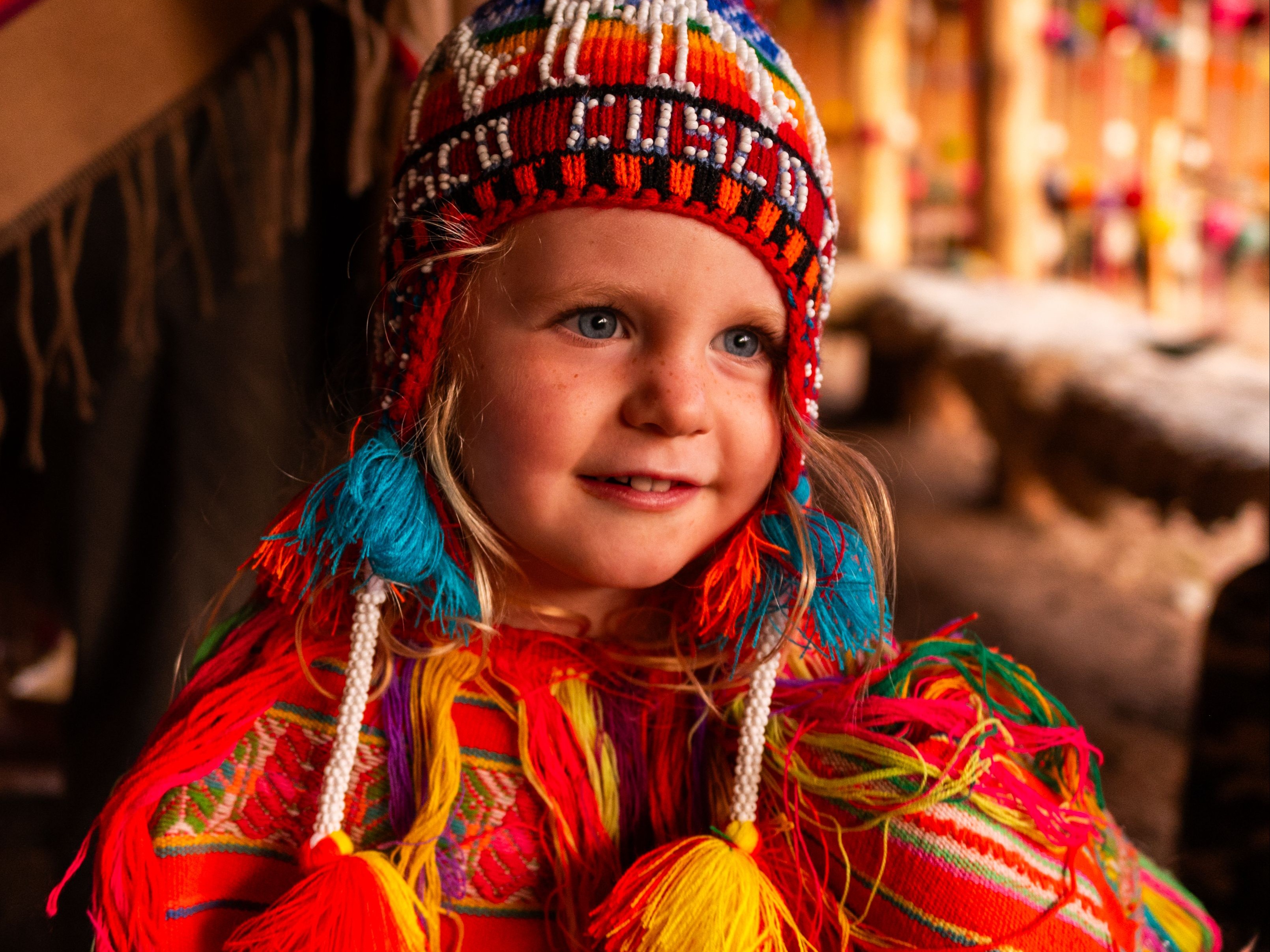 A person in a colorful traditional outfit with a beaded headpiece stands in front of a blurred indoor setting with wooden elements.