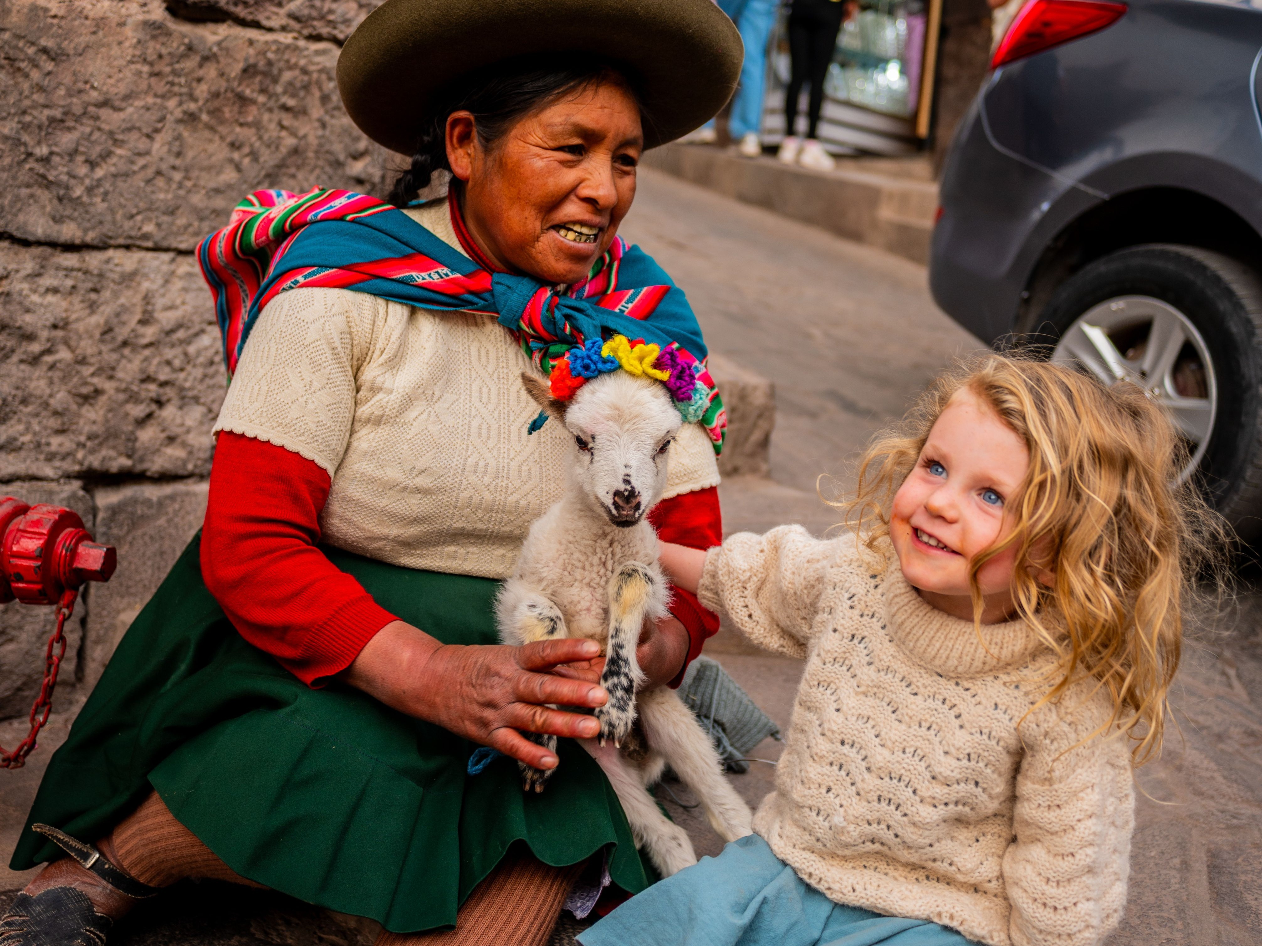 A person in traditional Andean clothing sits with a child and a small llama, surrounded by onlookers.