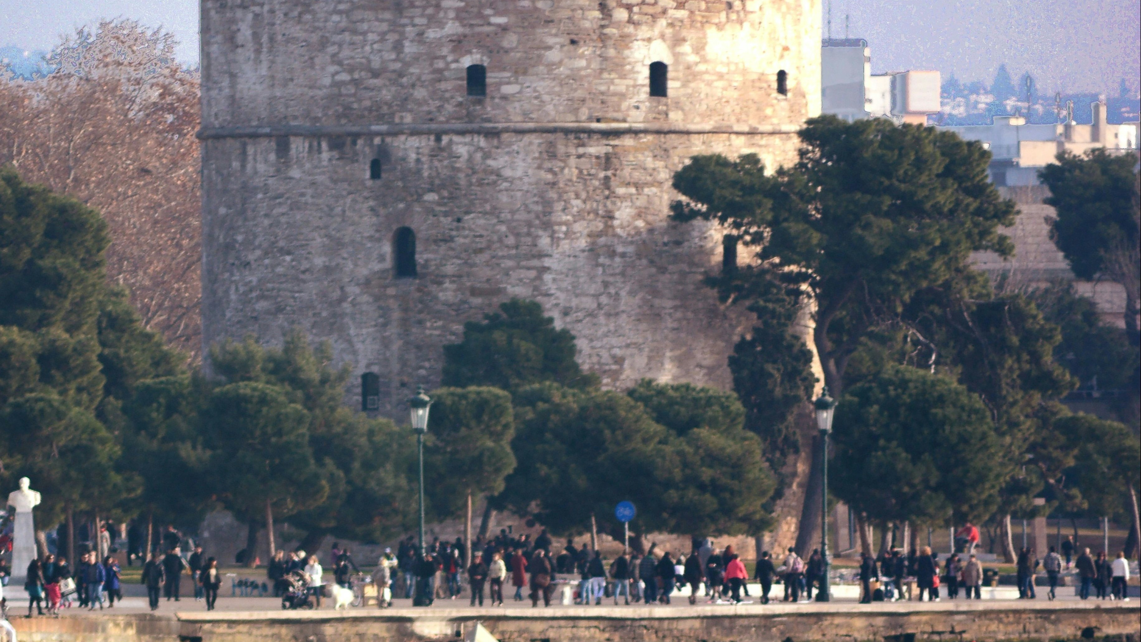 A large stone tower with battlements is reflected in the water, with people and trees at its base and a small boat nearby.