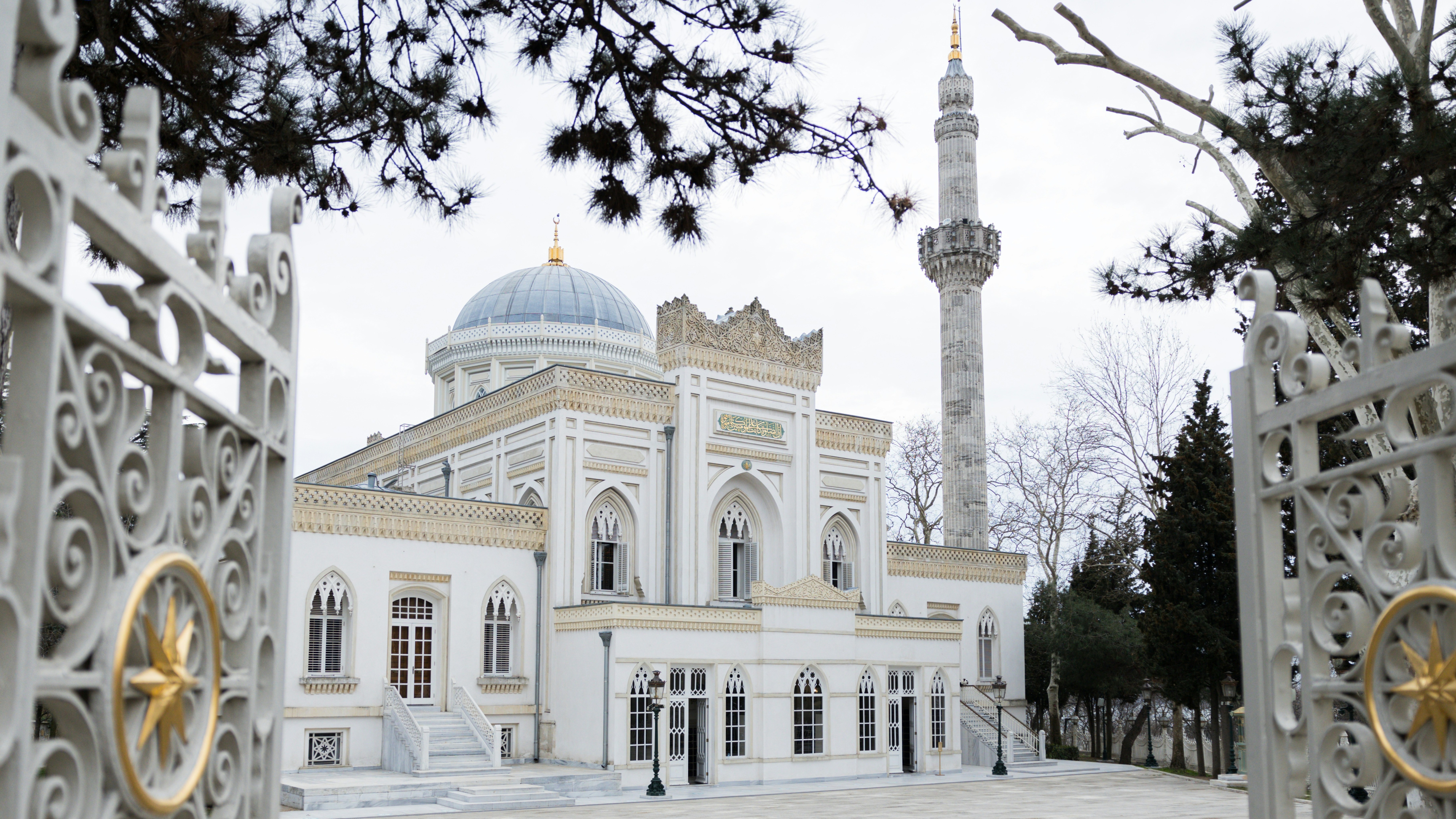 An ornate white building with a dome and minaret is viewed through open gates adorned with golden stars.