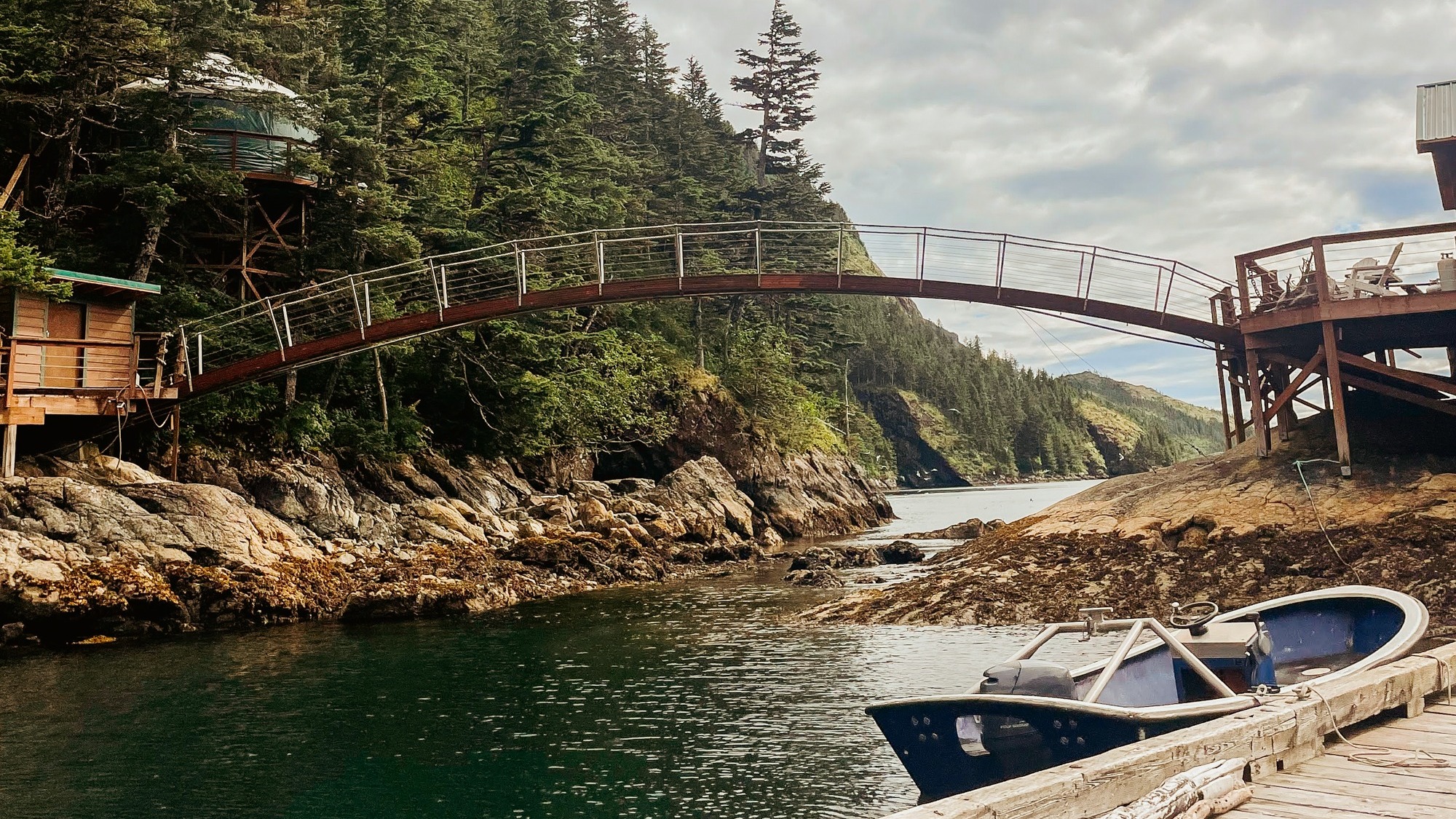 A serene wooden bridge spans over water, connecting rocky outcrops, a boat docked nearby and forested hills in the background.