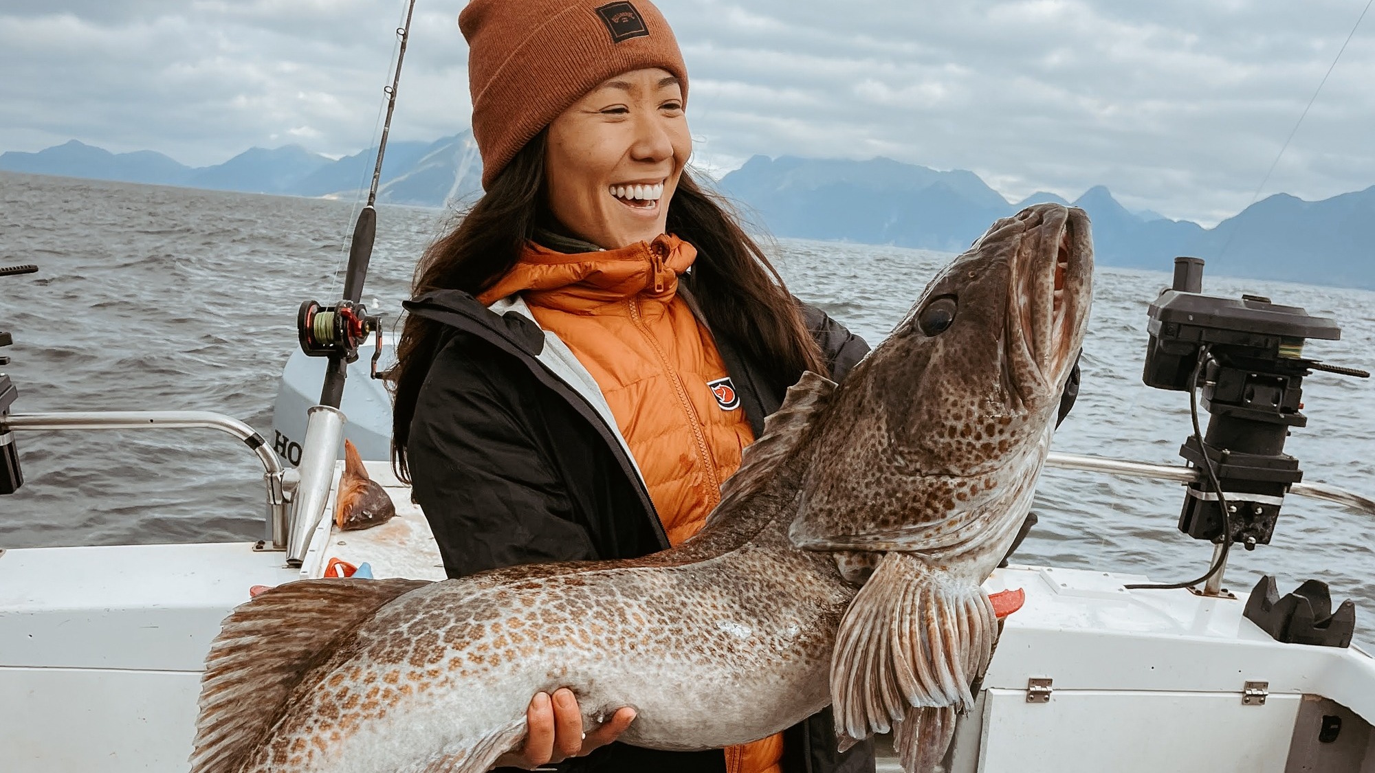 An individual on a boat holding a large fish, with mountains and overcast skies in the background.