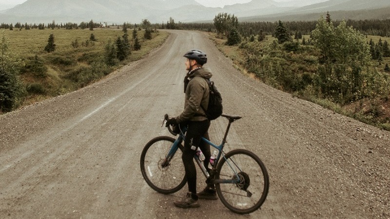 An individual with a bicycle standing on a dirt road, surrounded by a vast landscape of trees and mountains.