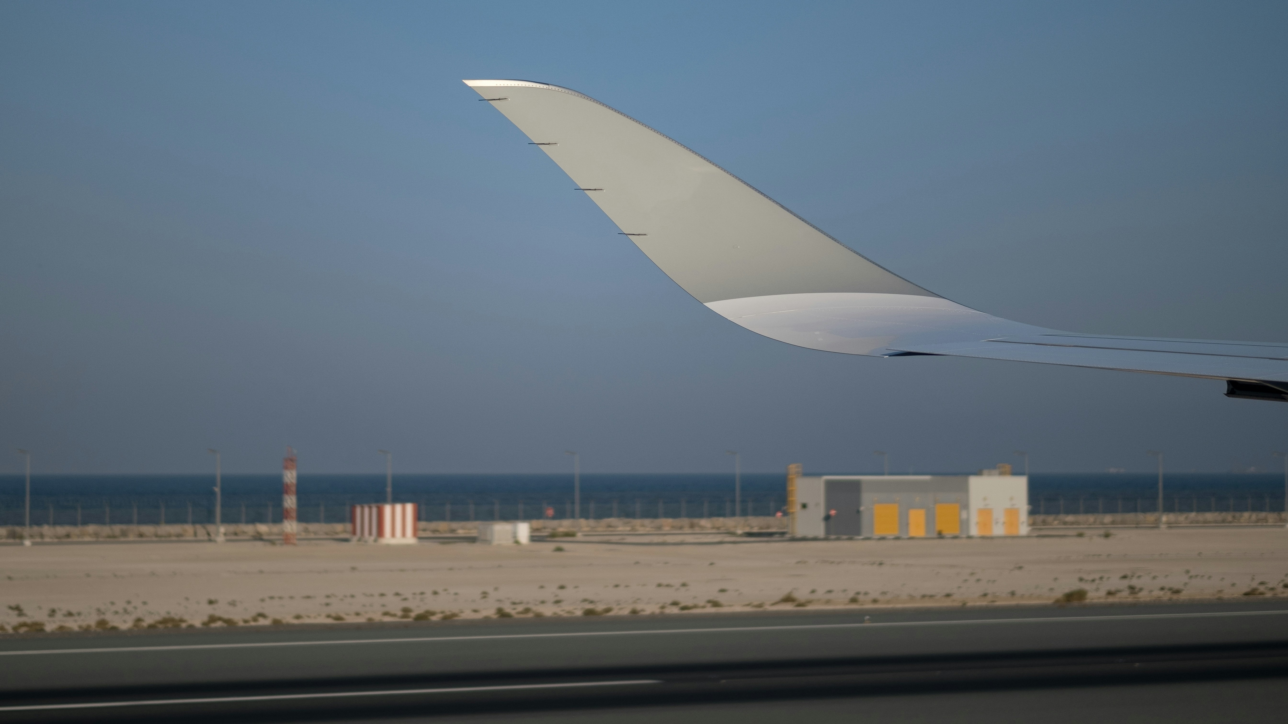 The image shows an airplane's wingtip against a clear sky, sandy ground and airport infrastructure.