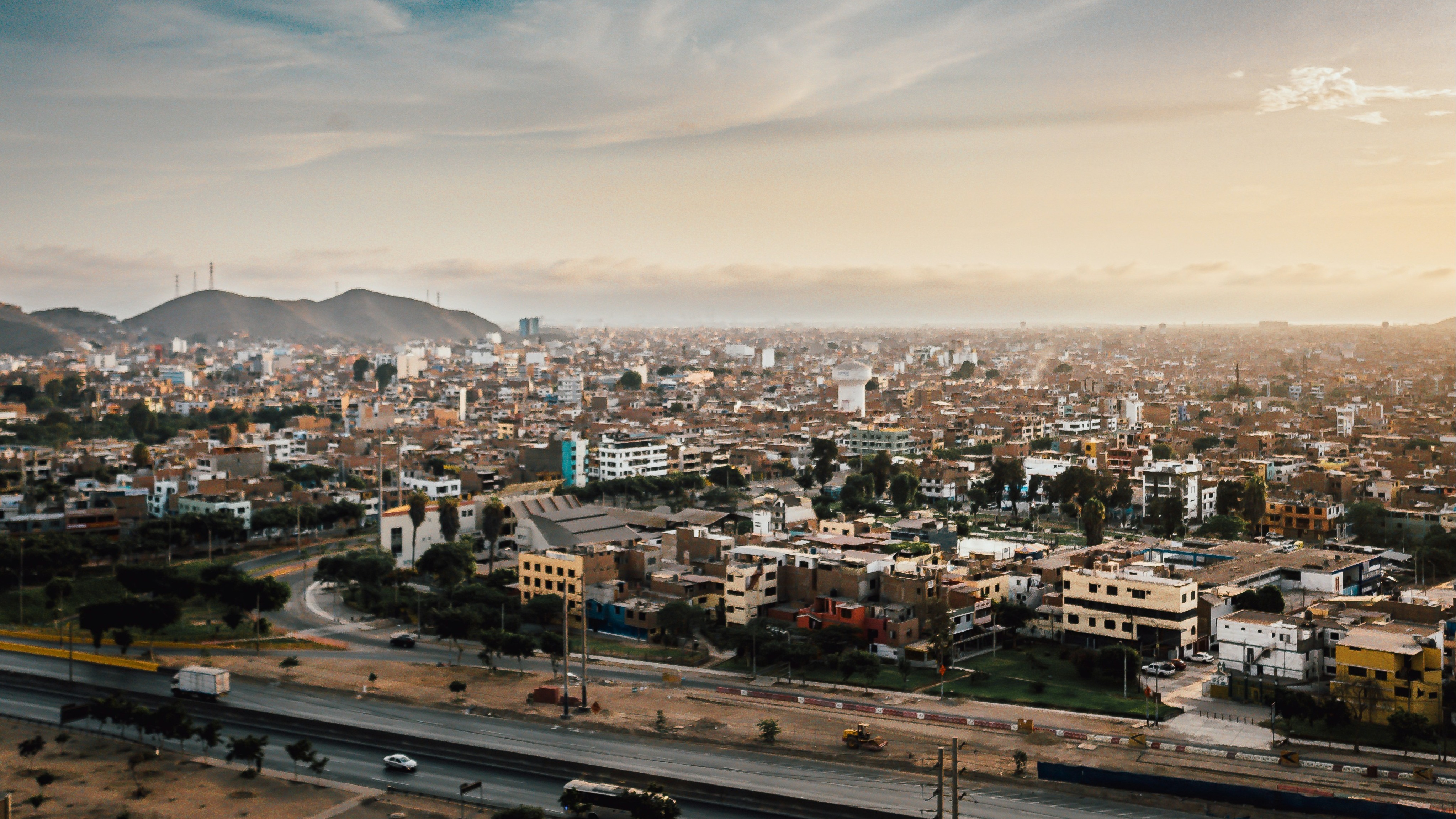 An aerial view of a cityscape during sunset, with buildings, roads and mountains under a partly cloudy sky.