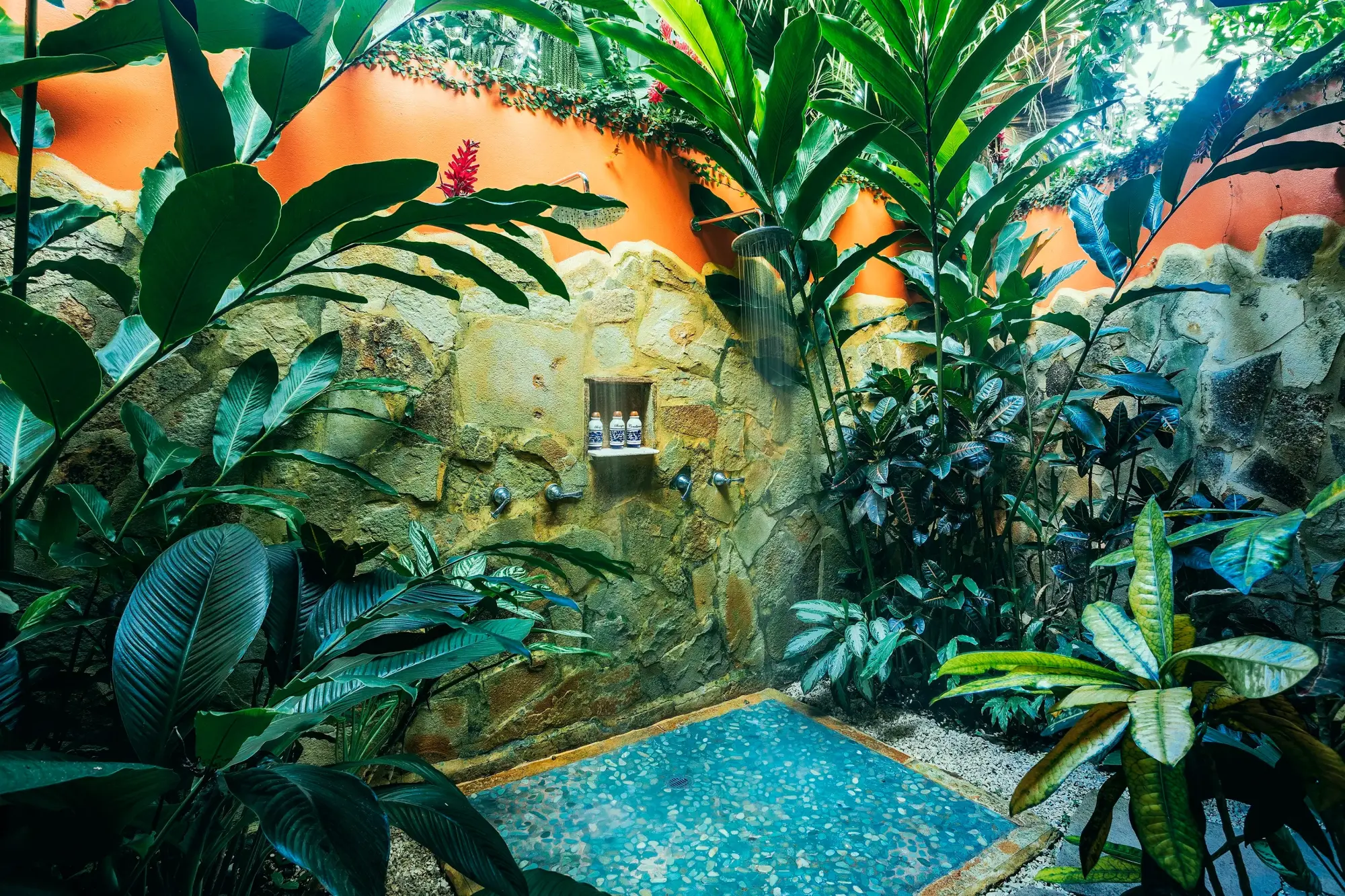 Outdoor shower surrounded by plants and stone walls; a tile floor catches water from the rain-like shower head