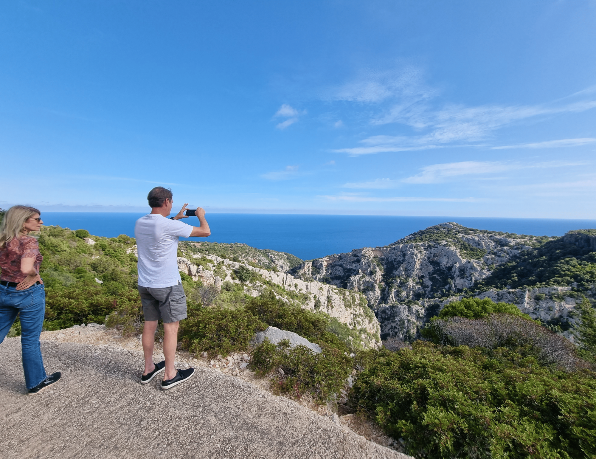 Two people standing high up on a lookout, take pictures of mountains and the ocean below on a sunny day.