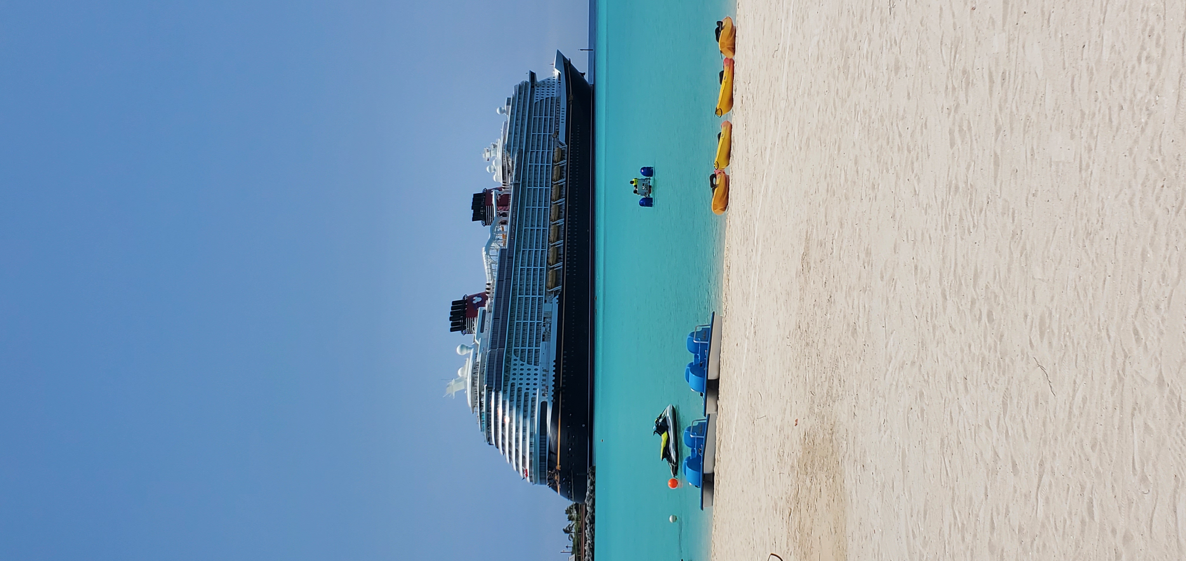 A cruise ship docks in the harbor across from a sandy beach on a clear day.