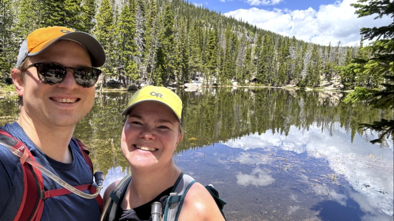 Two individuals take a selfie in front of a scenic forest and mountain lake, with reflections visible on the water’s surface.