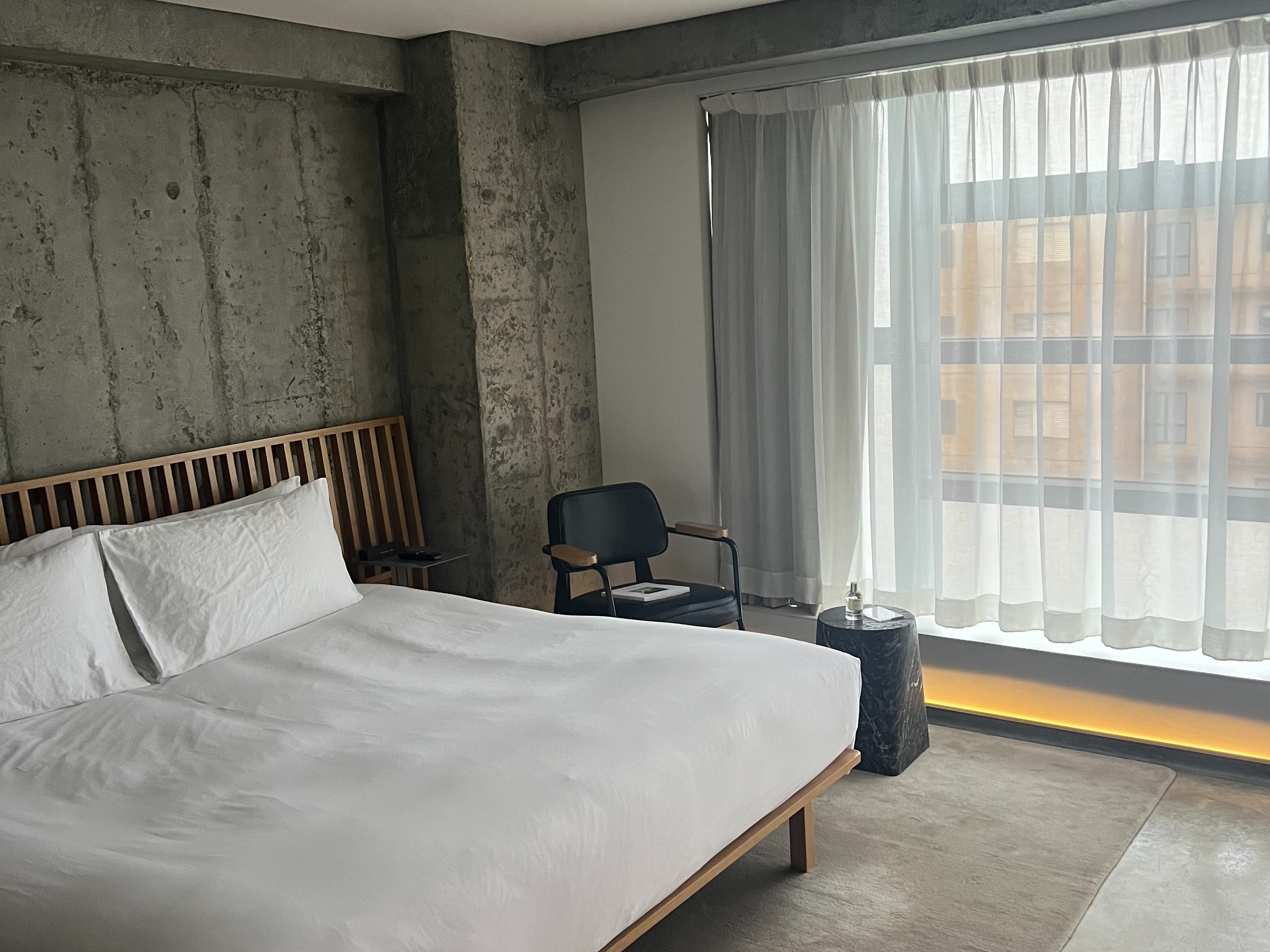 A modern hotel room with a bed, side table, lamp, cement walls and a window with sheer curtains in a clean, minimalistic design.