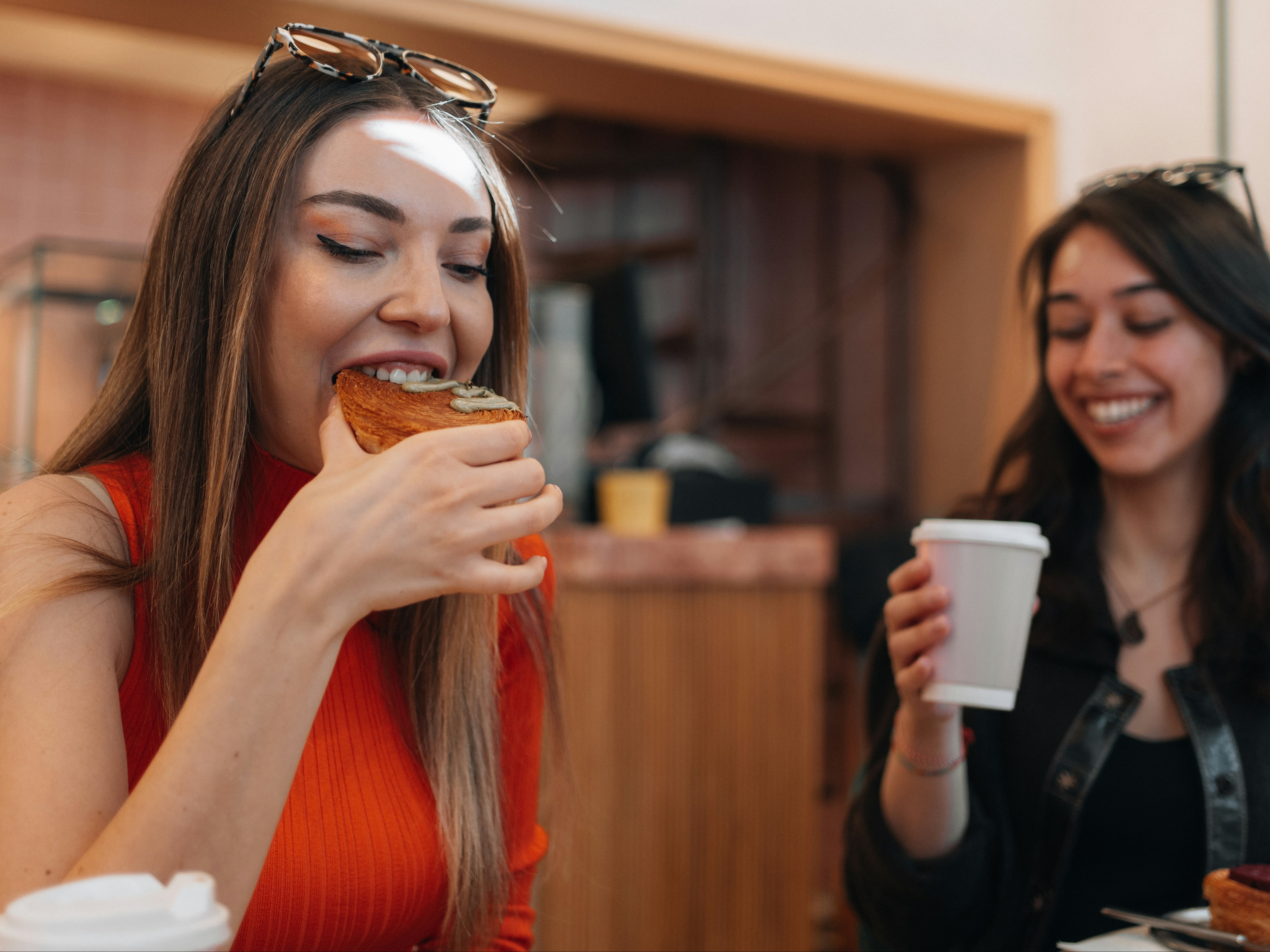 Two women smiling over coffee and a pastry, one woman is taking a bite.