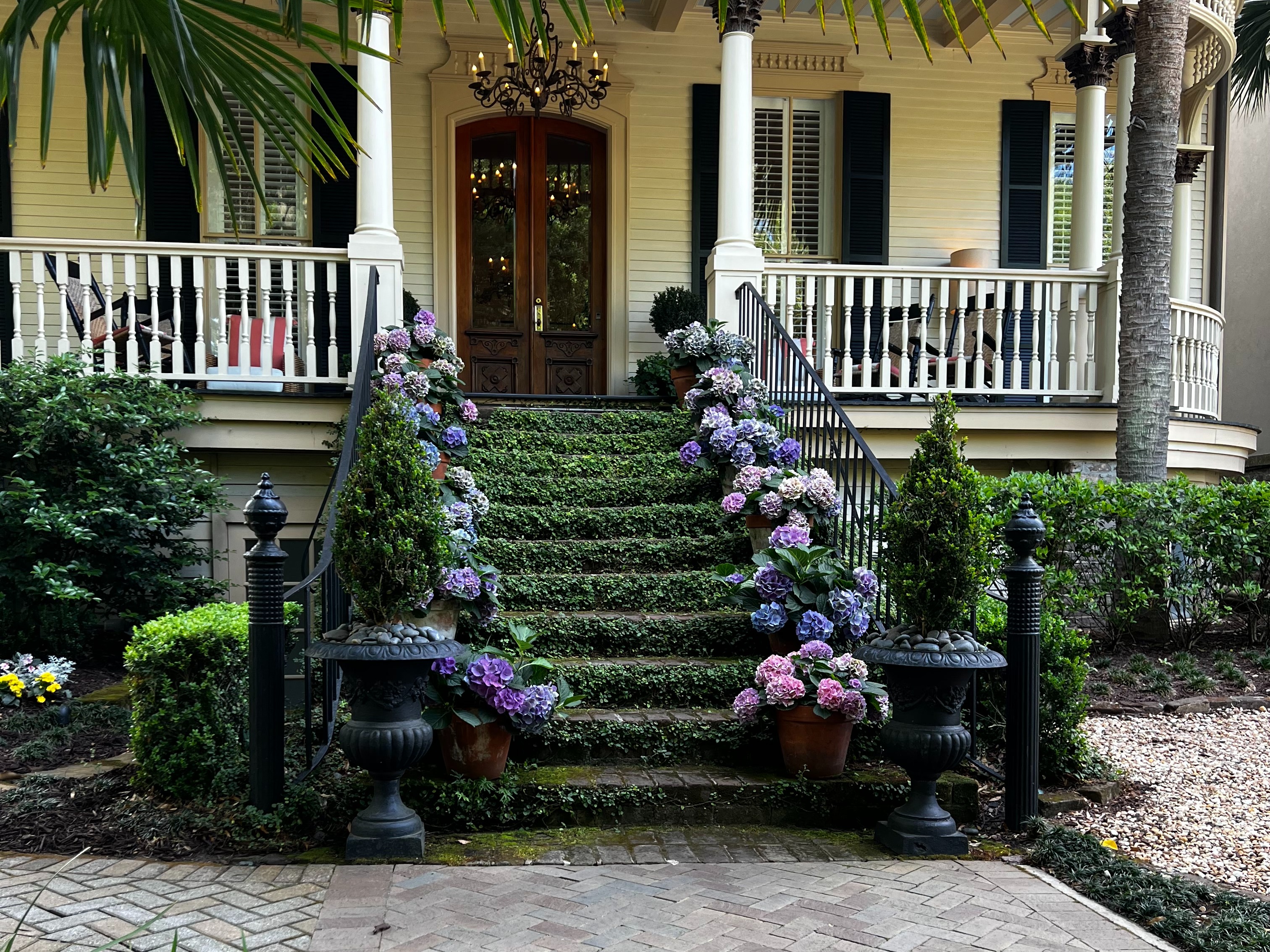 A view of the front porch of a beautiful home with white pillars, railing and steps covered in green moss and purple flowers lining the staircase.