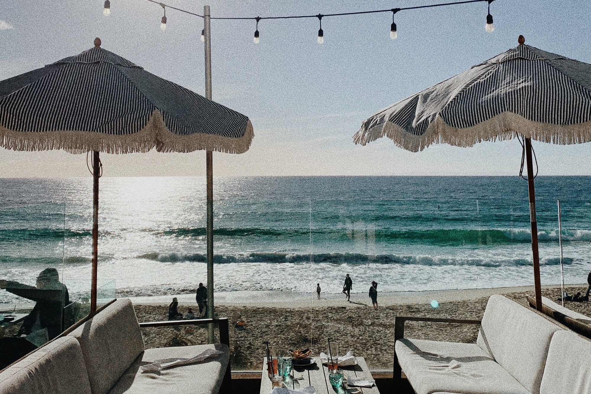 Outdoor seating at a restaurant with white couches, a table and pinstriped fringe umbrellas overlooking the beach on a sunny day.