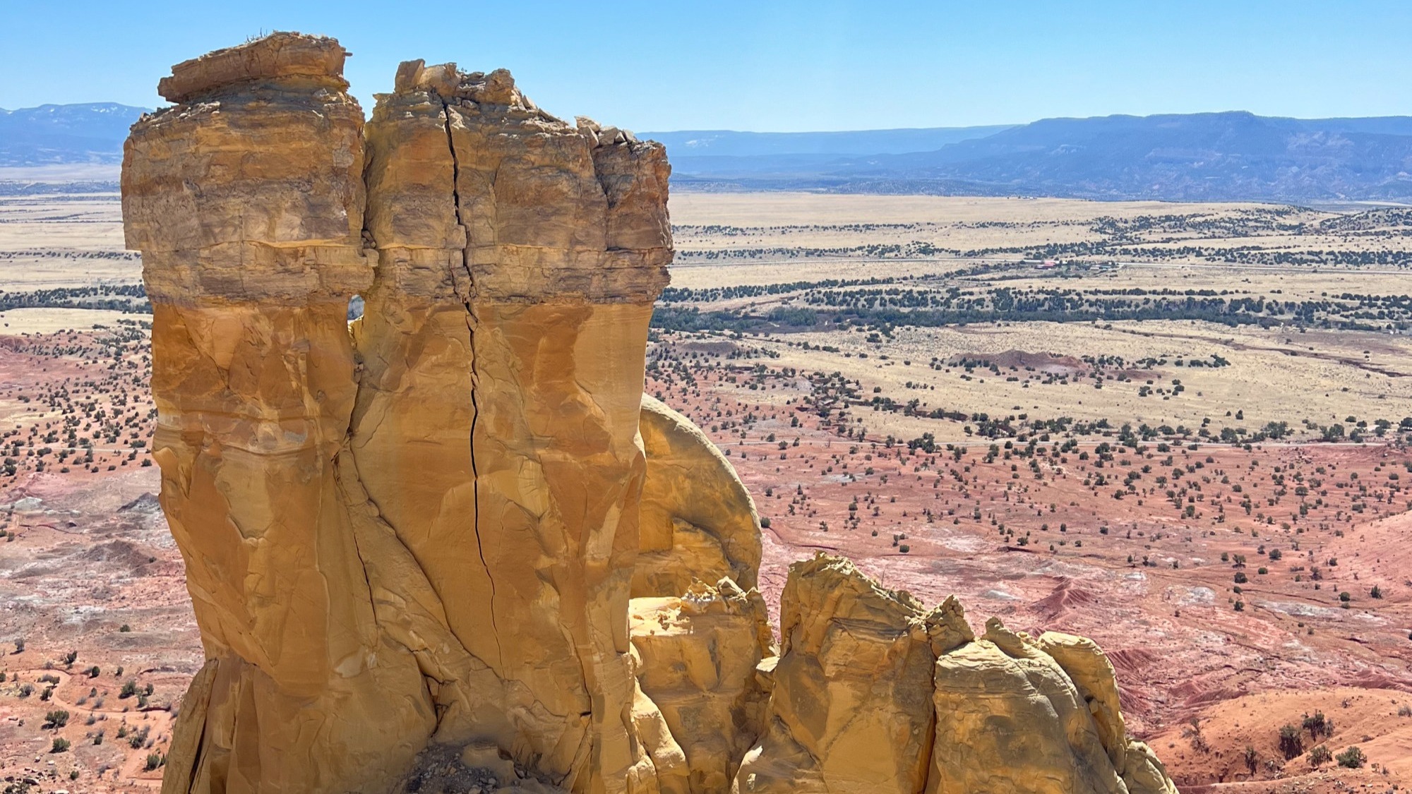 A spire of desert rocks with red desert surrounding and mountains in the distance.