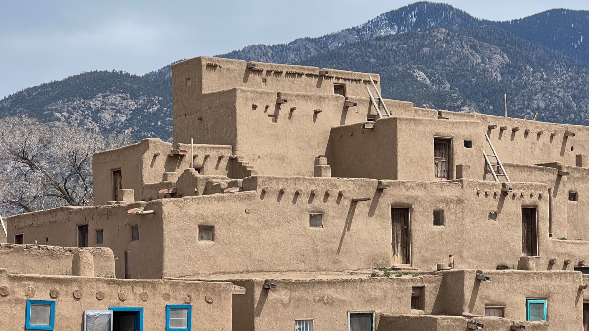A complex adobe building with several stories and rooms with mountains in the background.