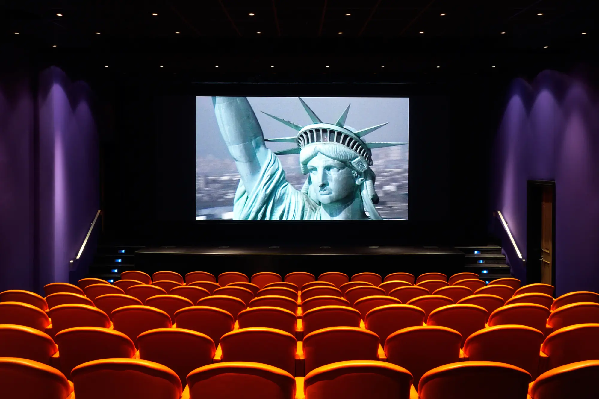 Rows of orange seats descend, culminating in a movie theater screen depicting a closeup of the Statue of Liberty's face