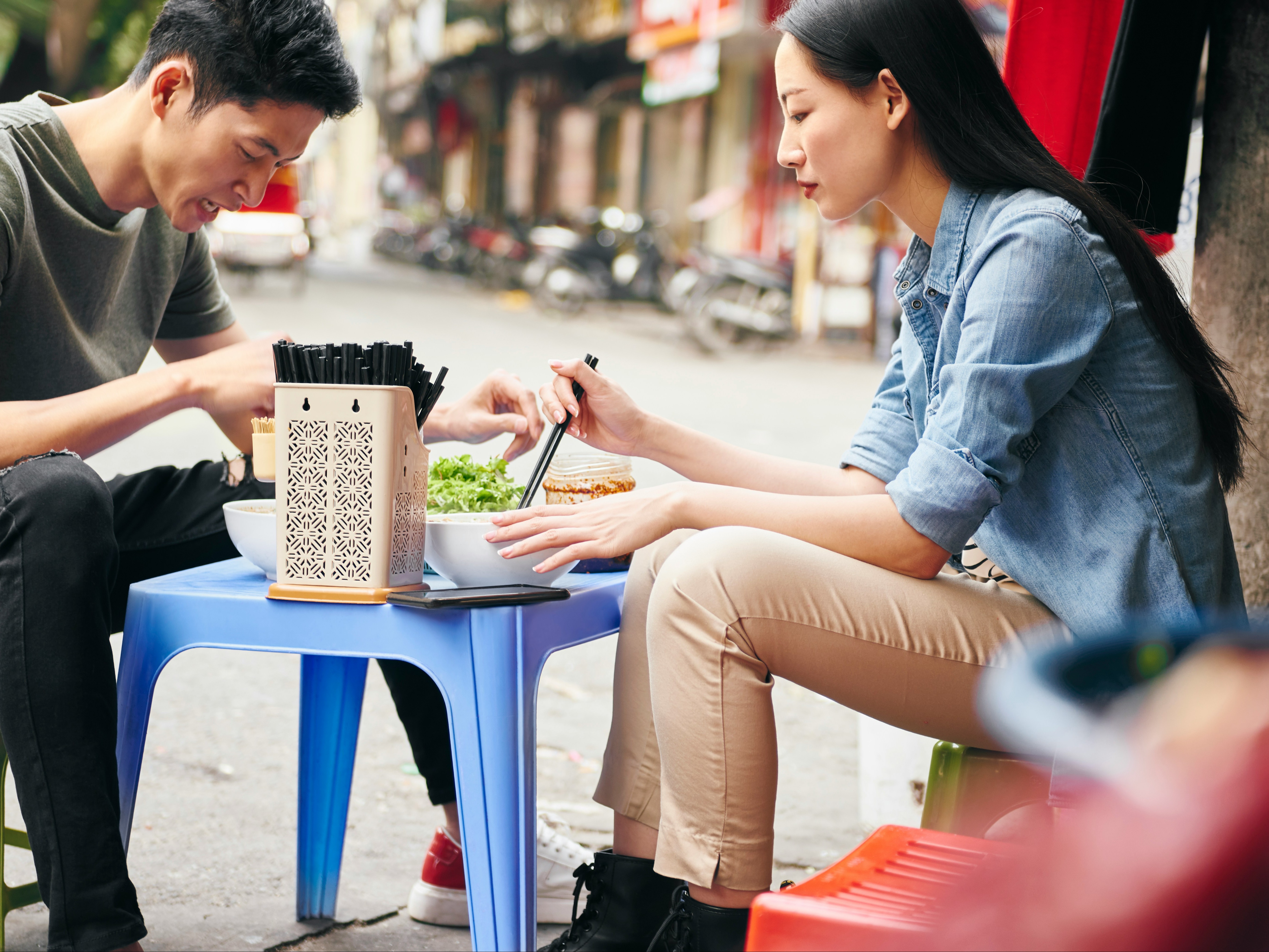 Two people sitting at a short table outside in a street eating food