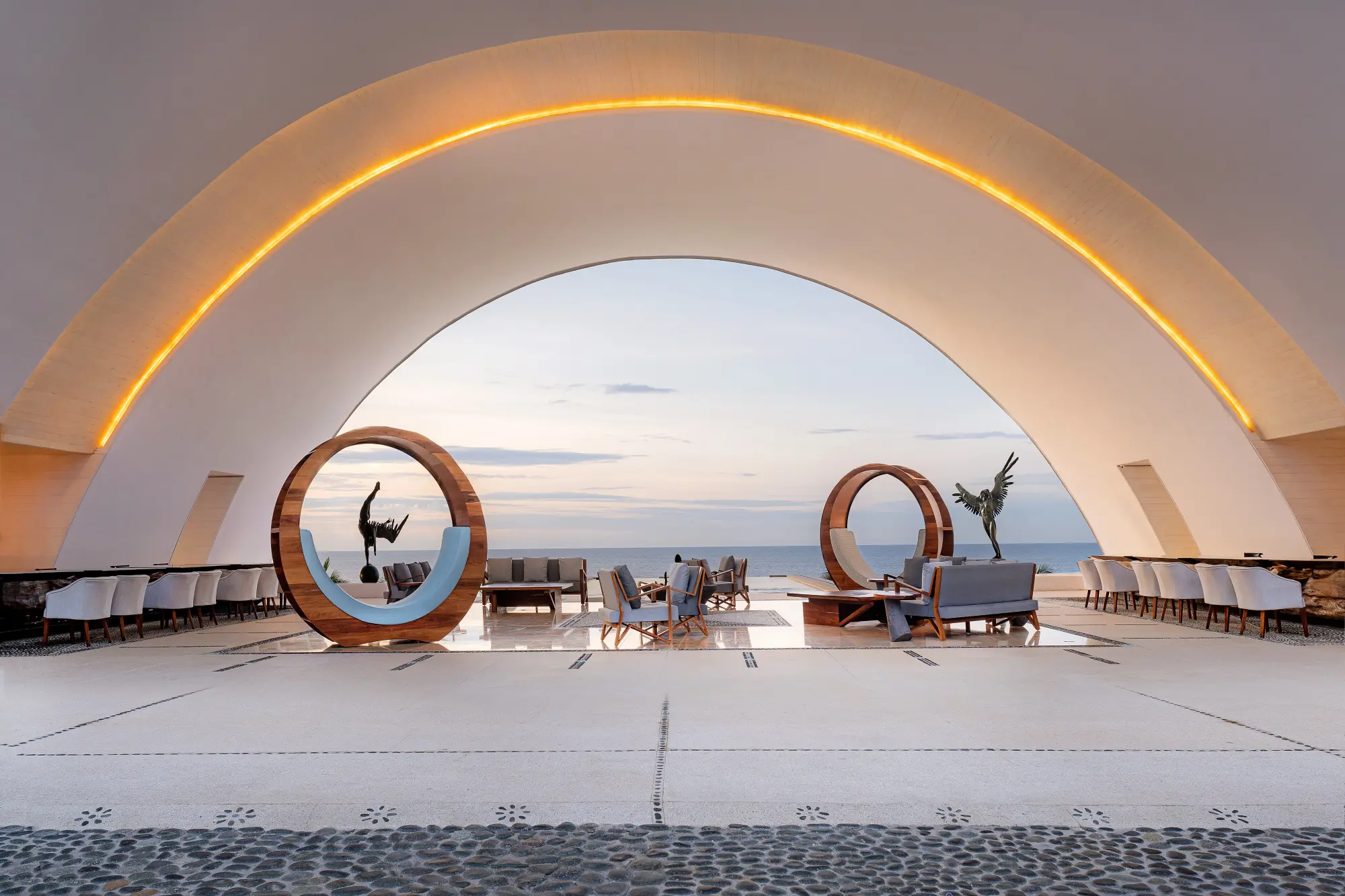 A chic space with elegant statues and unique, contemporary seating arrangements all underneath a well-lit semi-dome with ocean views