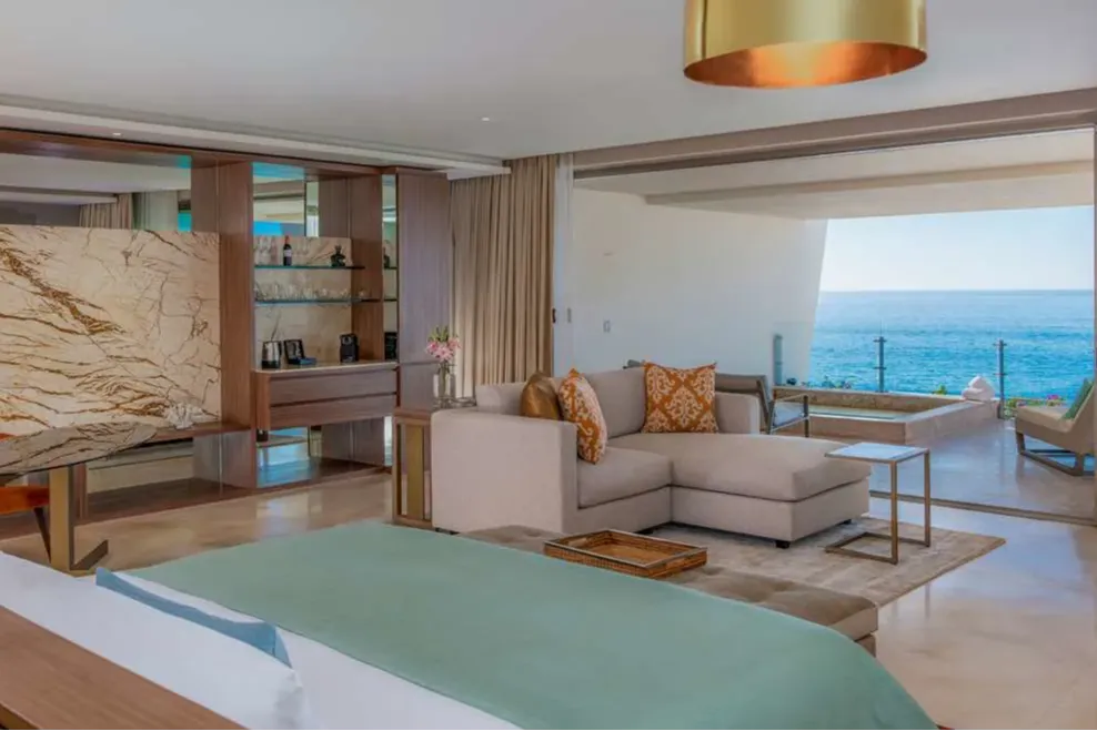 A large bed with pastel bedding leads to a comfortable seating area that, in turn, leads to a glass-walled balcony with ocean views