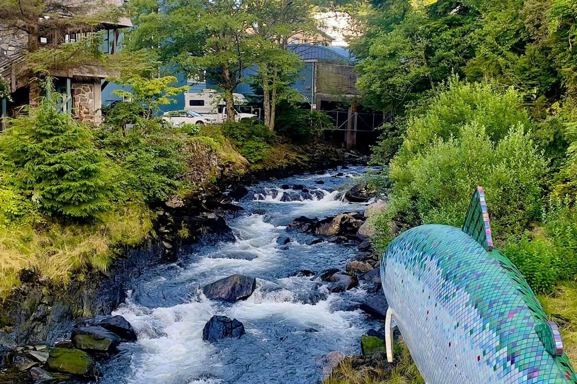 A view of a creek rushing down between a lush forest with buildings along one side and a fish sculpture in the foreground.