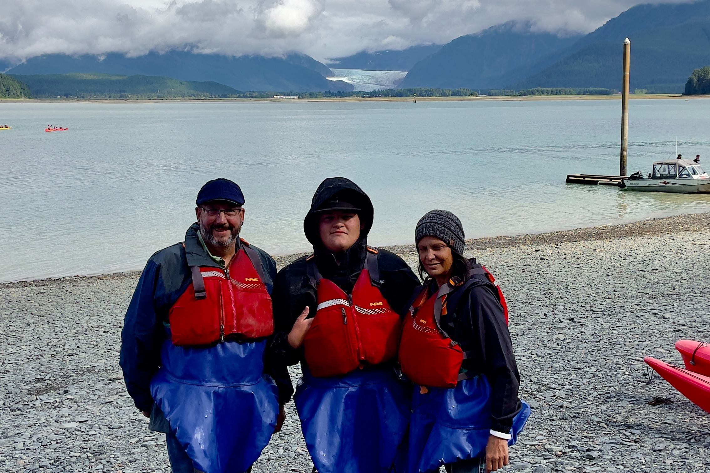 The advisor and her family geared up in warm clothing and life jackets for Mendenhall Lake kayaking adventure with the water and mountains behind them on an overcast day.