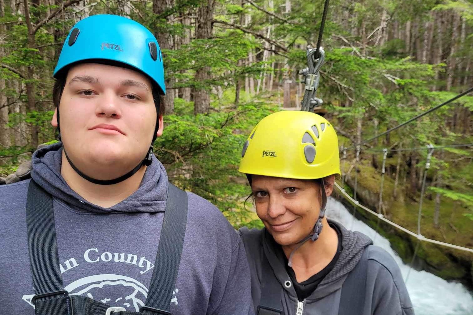 Advisor and a boy pose wearing hard hats with a zipline and a forest behind them at Grizzly Falls, Skagway, Alaska.