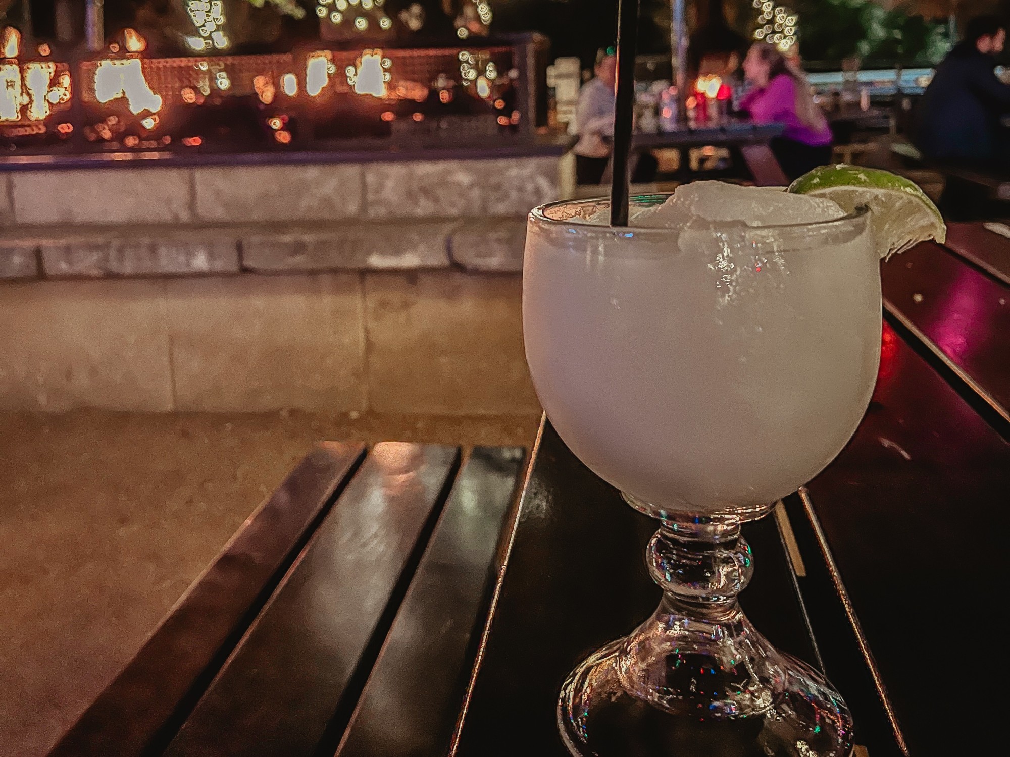 A blended cocktail with a lime garnish on a wooden table with a fire in the background.